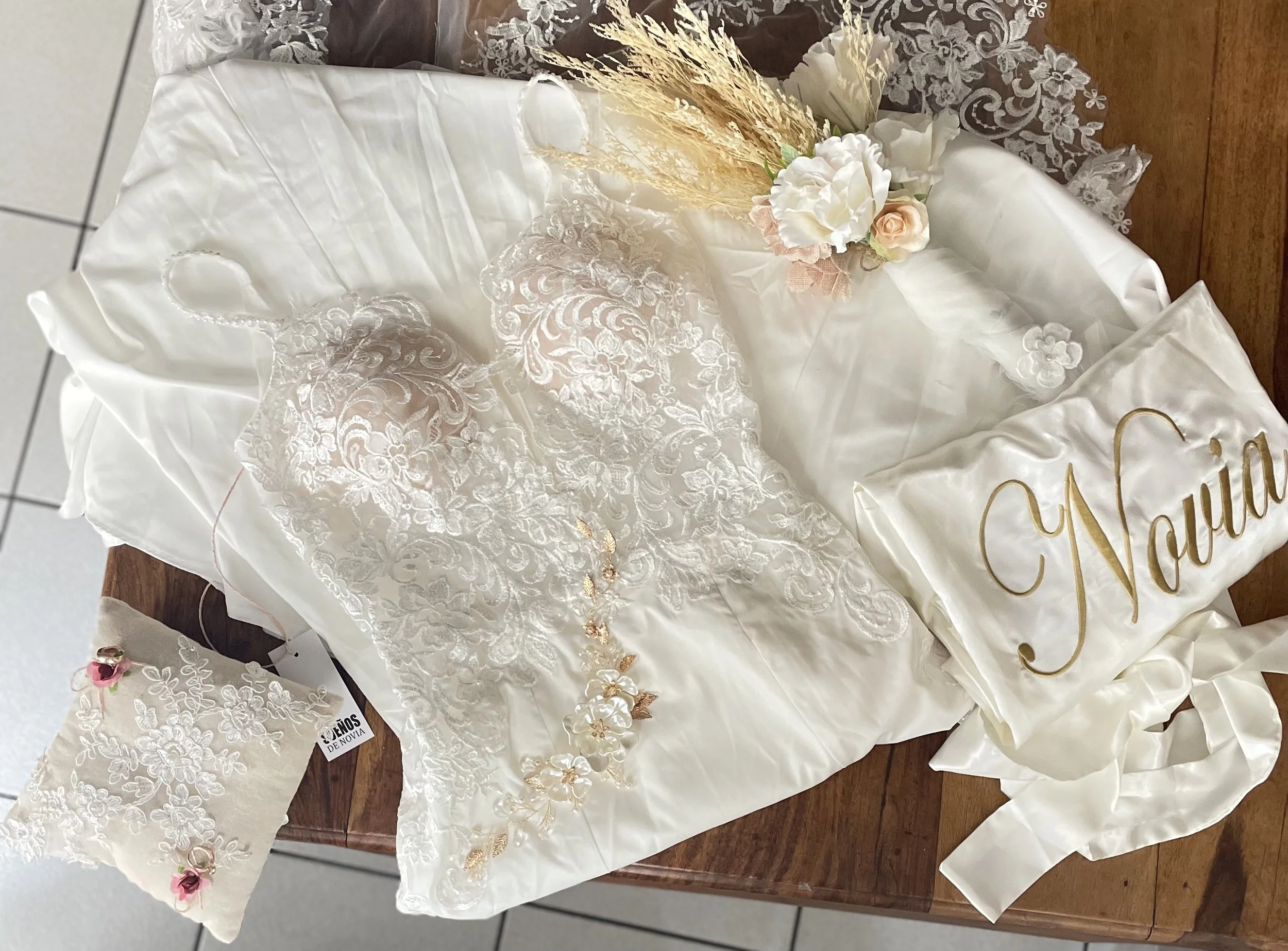 Robe de mariée en dentelle blanche avec un bouquet de fleurs séchées et un coussin d'alliances, posés sur une table en bois.