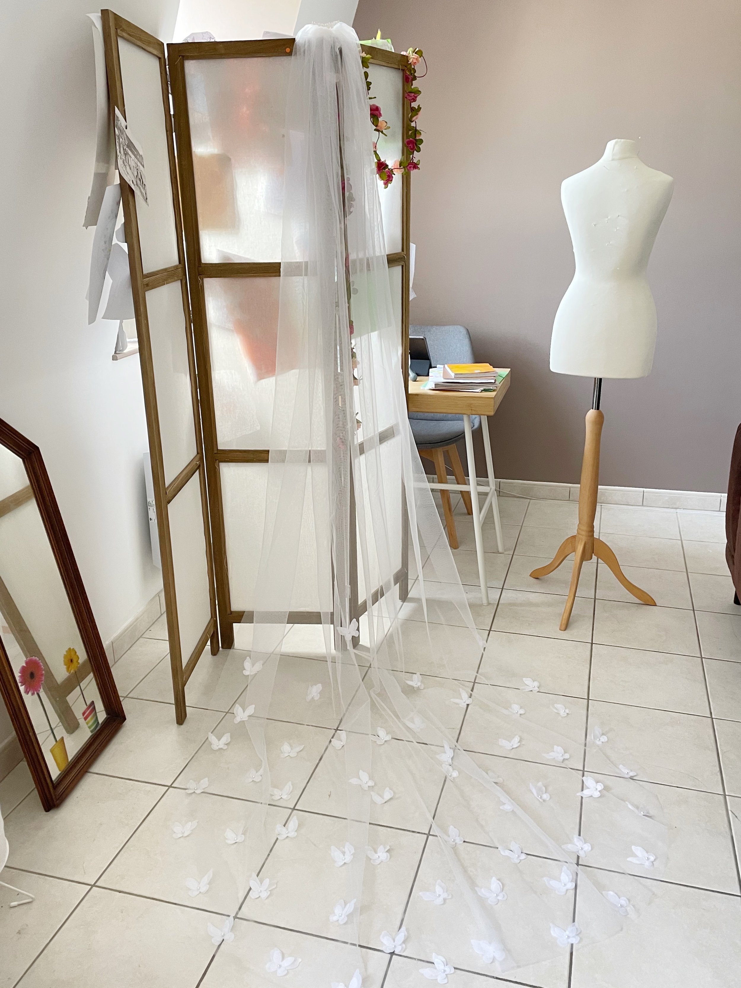 Voile de mariée avec décorations de papillons blancs sur le sol, mannequin, paravent en bois, miroir incliné et table avec livres dans une pièce bien éclairée.