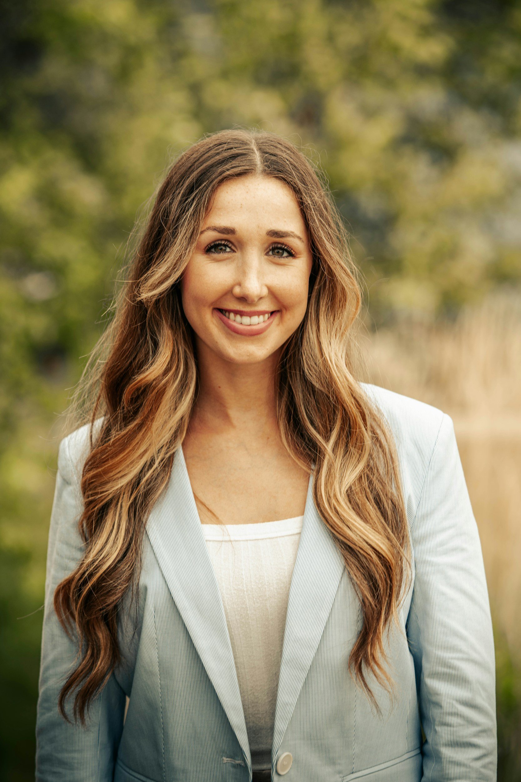 A smiling woman with long wavy hair, wearing a light-colored blazer and a white top, standing outdoors with a blurred green and yellow background.