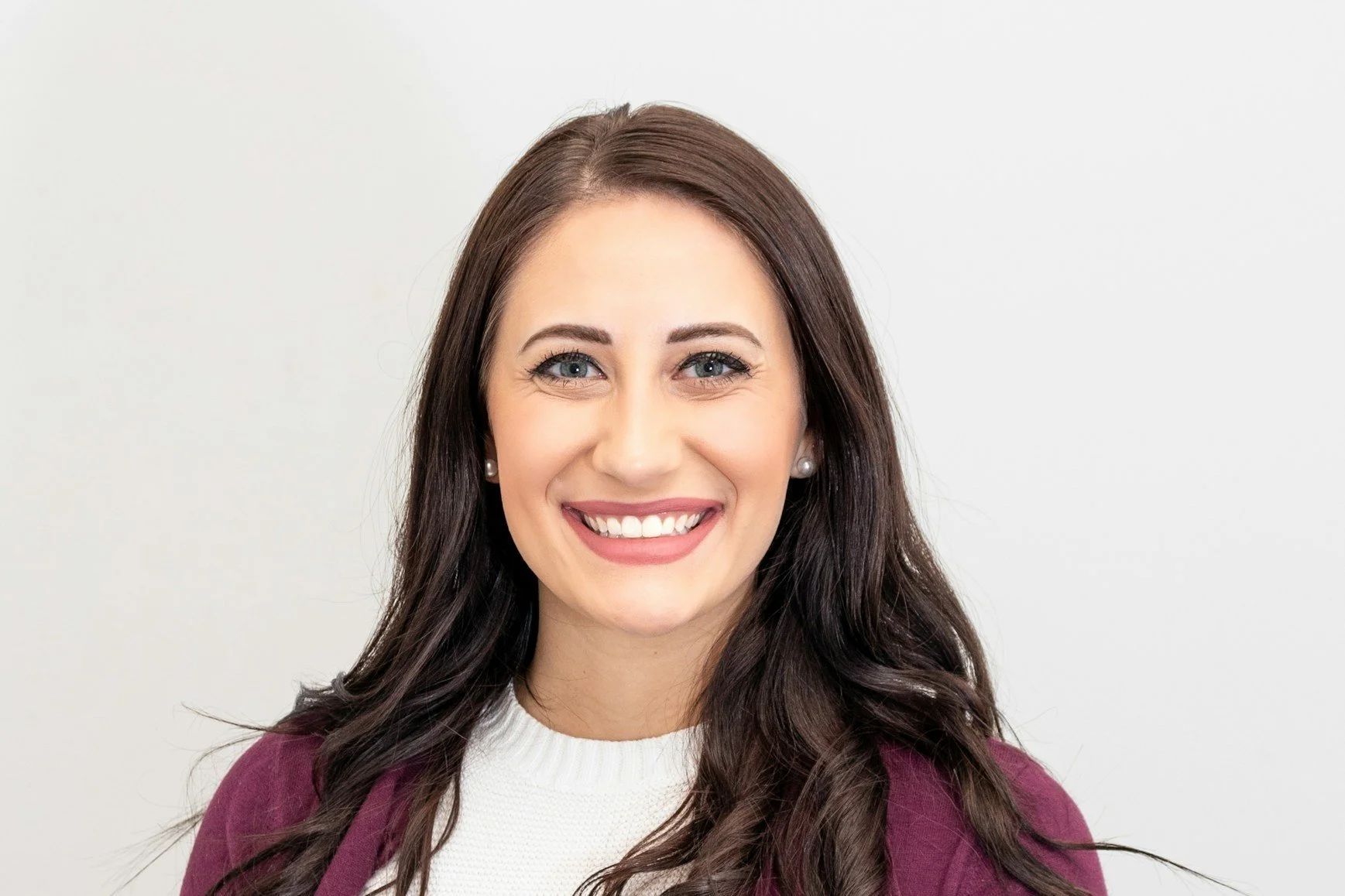 A woman with long brown hair, blue eyes, and a bright smile wearing a white top and a purple cardigan against a plain white background.