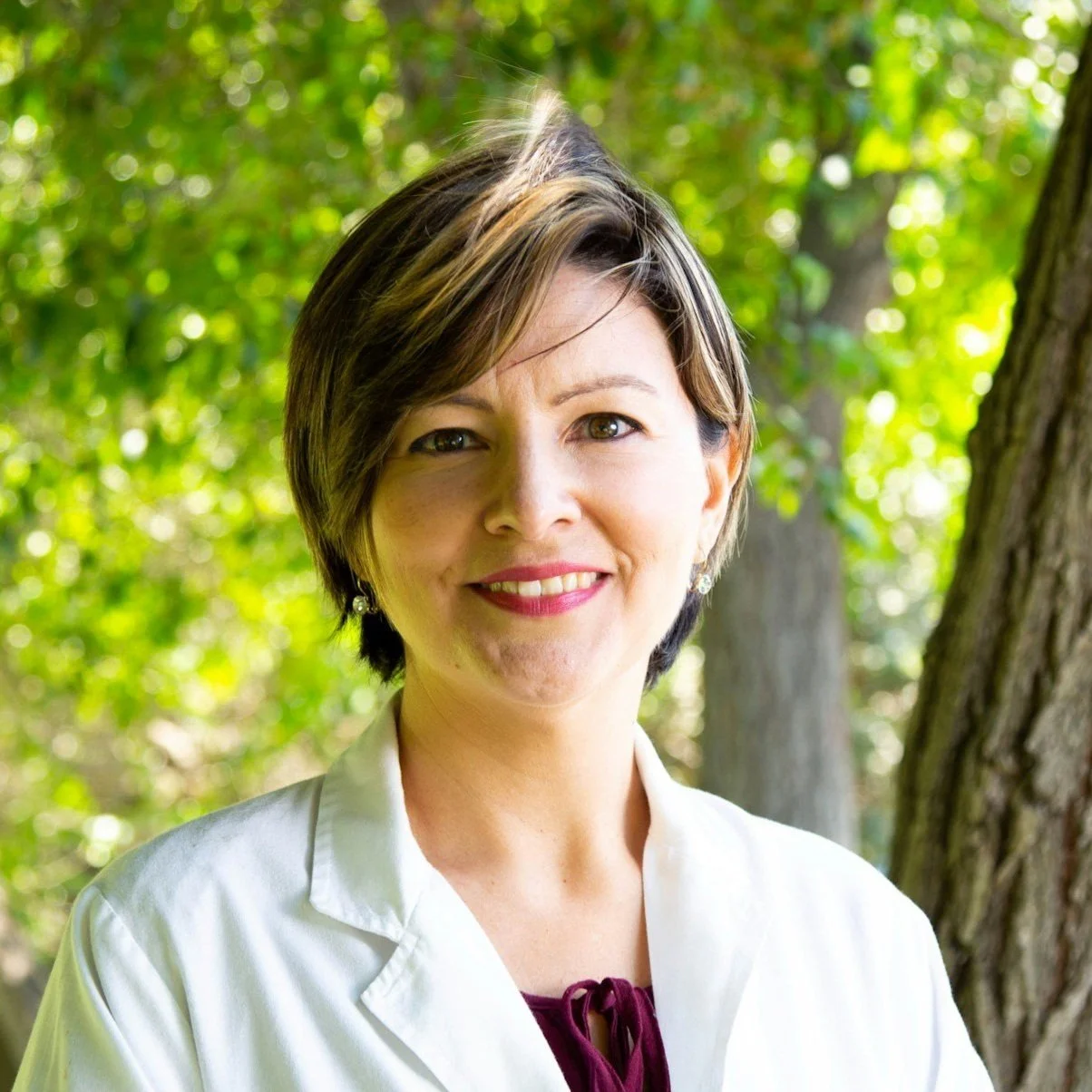 A woman with short diverse brown hair and light skin smiling outdoors with green trees in the background.