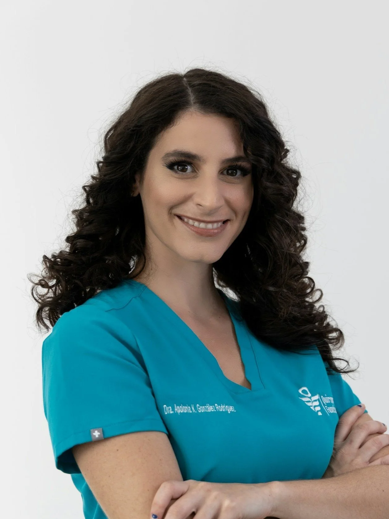 A woman with long, curly brown hair and light skin wearing a teal medical scrub top with her arms crossed, smiling against a plain white background.