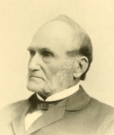 Historical black-and-white portrait of a stern-looking man with sideburns, wearing a suit and bow tie.