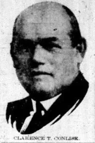 Black and white portrait of a man in formal attire, wearing a suit and tie, with short hair and glasses.