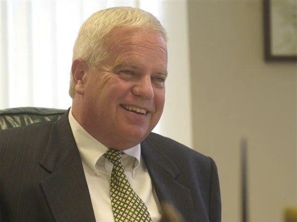 A smiling middle-aged man with light-colored hair, wearing a dark suit, white shirt, and patterned tie, sitting in an office.