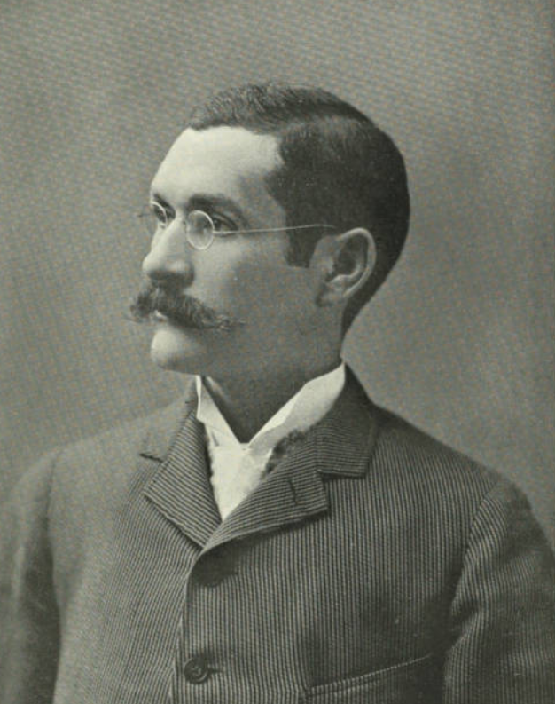 Black and white portrait of a man with a mustache, glasses, wearing a suit and a white shirt, facing left.