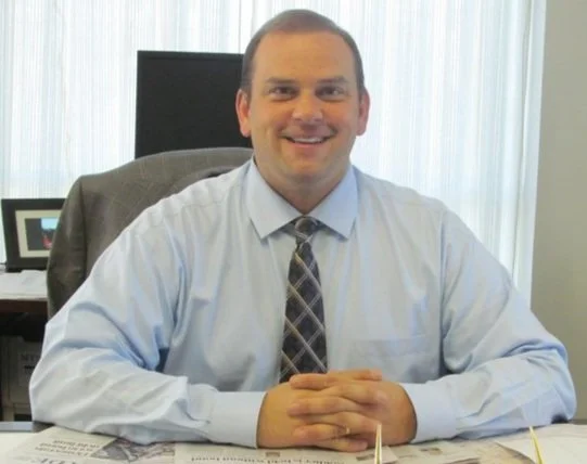 A man sitting at a desk in an office, smiling, wearing a light blue dress shirt and a patterned tie.