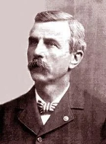 A black and white portrait of a man with a mustache, wearing a dark suit jacket and a bow tie, with short hair combed to the side.