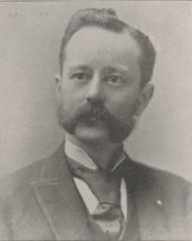 Black and white portrait of a man with a mustache, wearing a suit and bow tie, with a serious expression.
