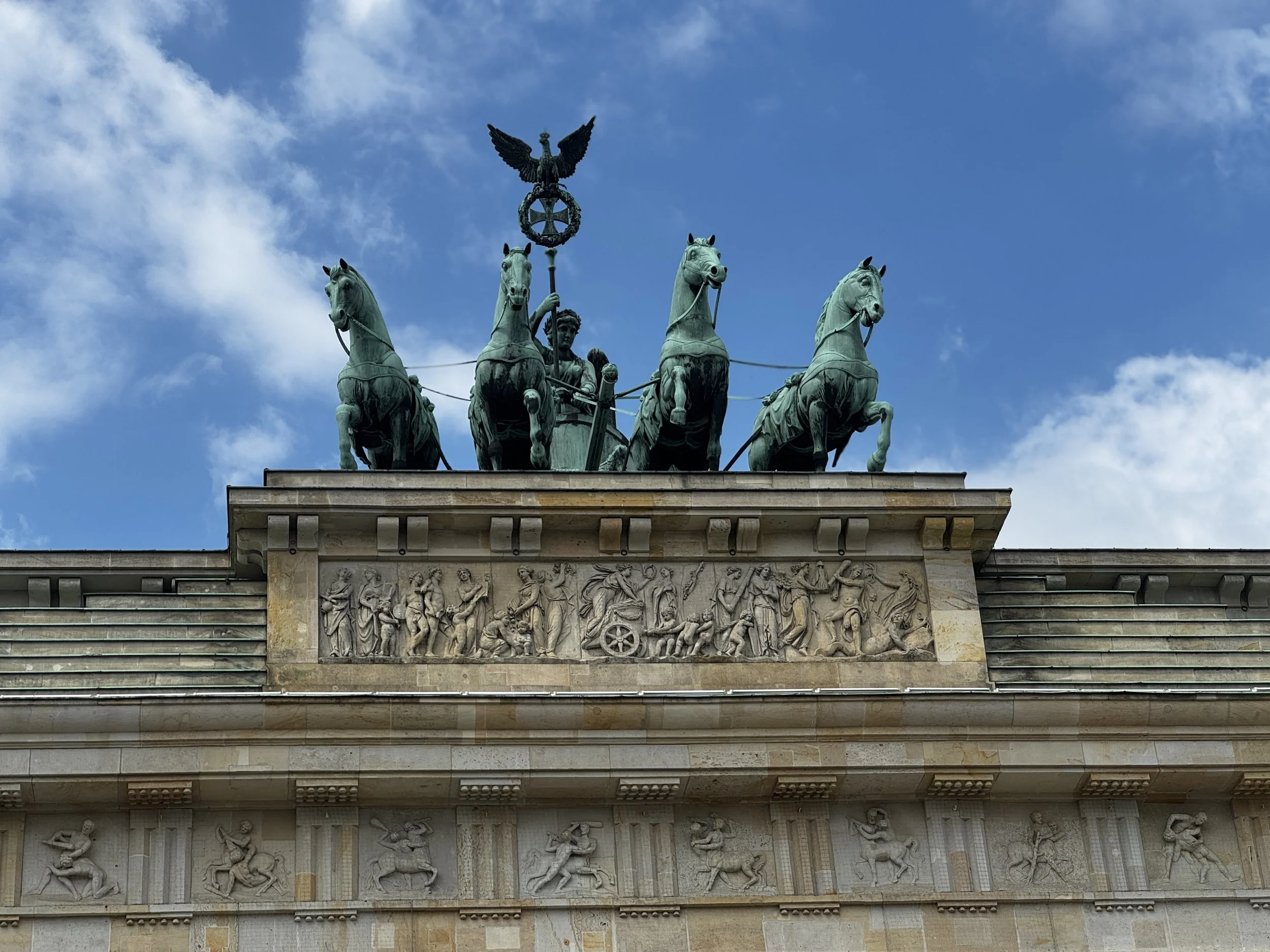 The Brandenburg Gate in Berlin, Germany, featuring a quadriga sculpture of a chariot drawn by four horses at the top, with historical relief sculptures on the lower part of the structure and a blue sky with some clouds background.