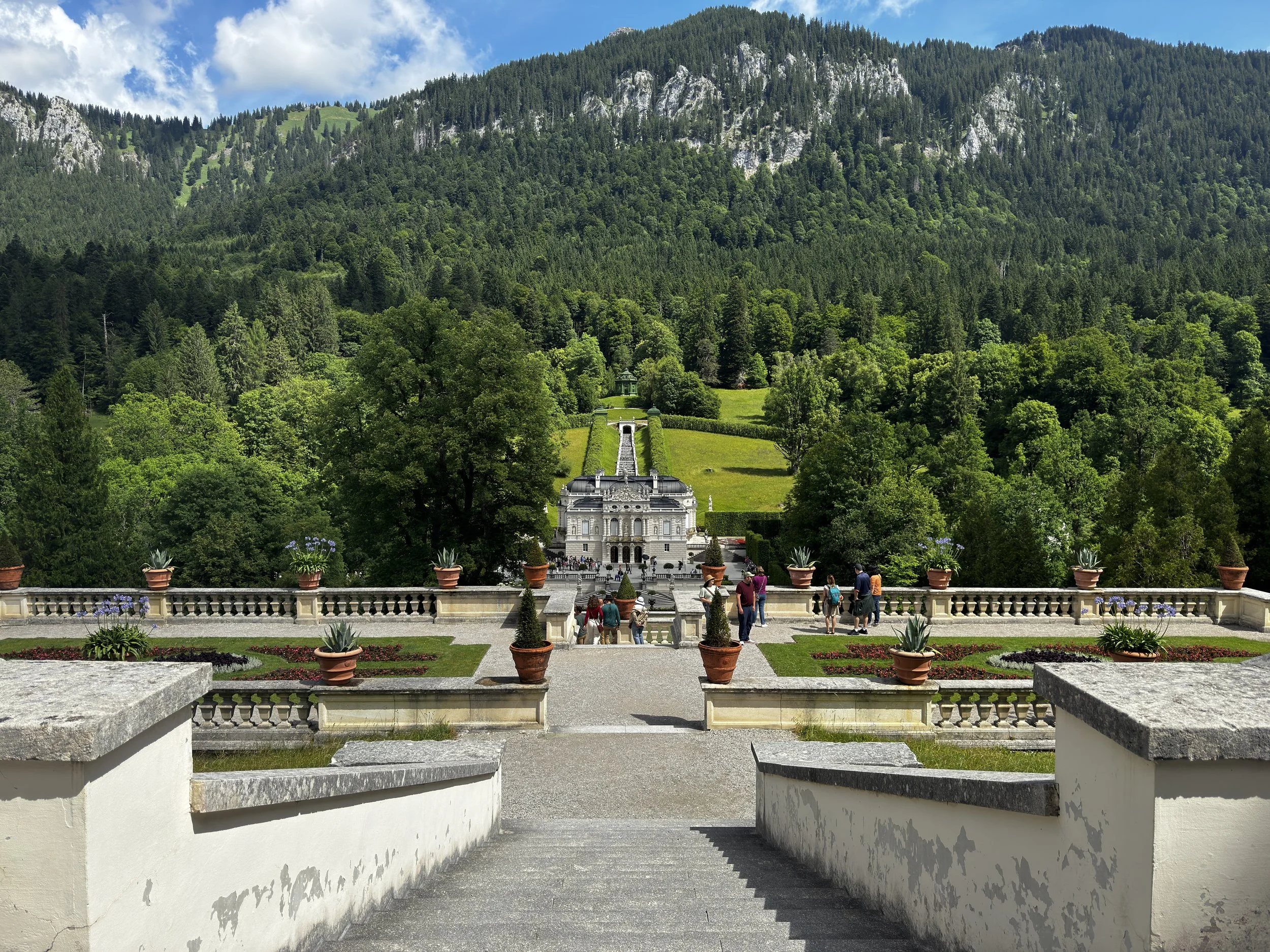 A grand terrace with potted plants and flower beds leading to a pathway, overlooking a lush green garden and a mountainside in the background.