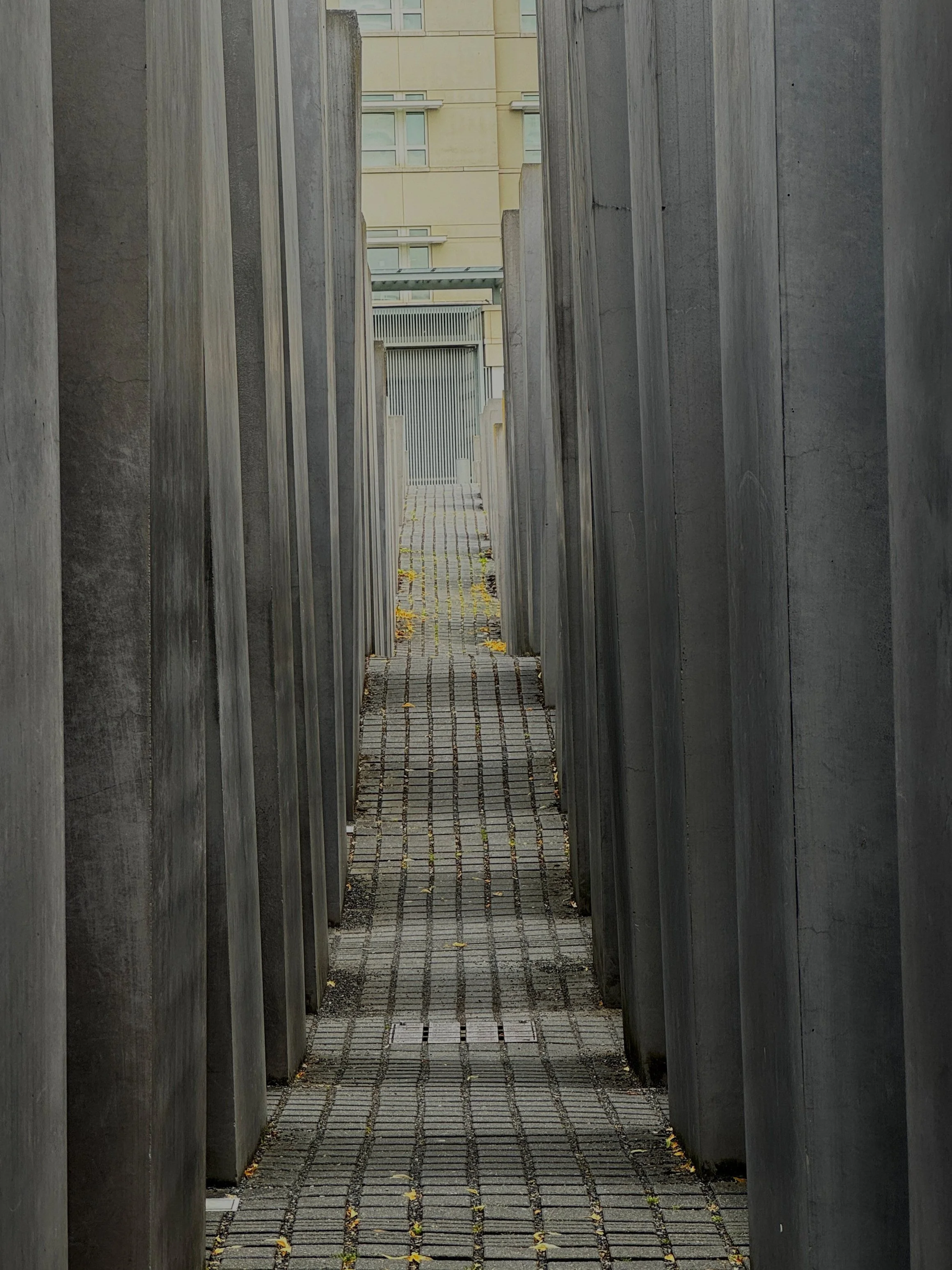 A narrow pathway between tall, dark stone monuments or memorials leading to a gated area with a modern residential building in the background.