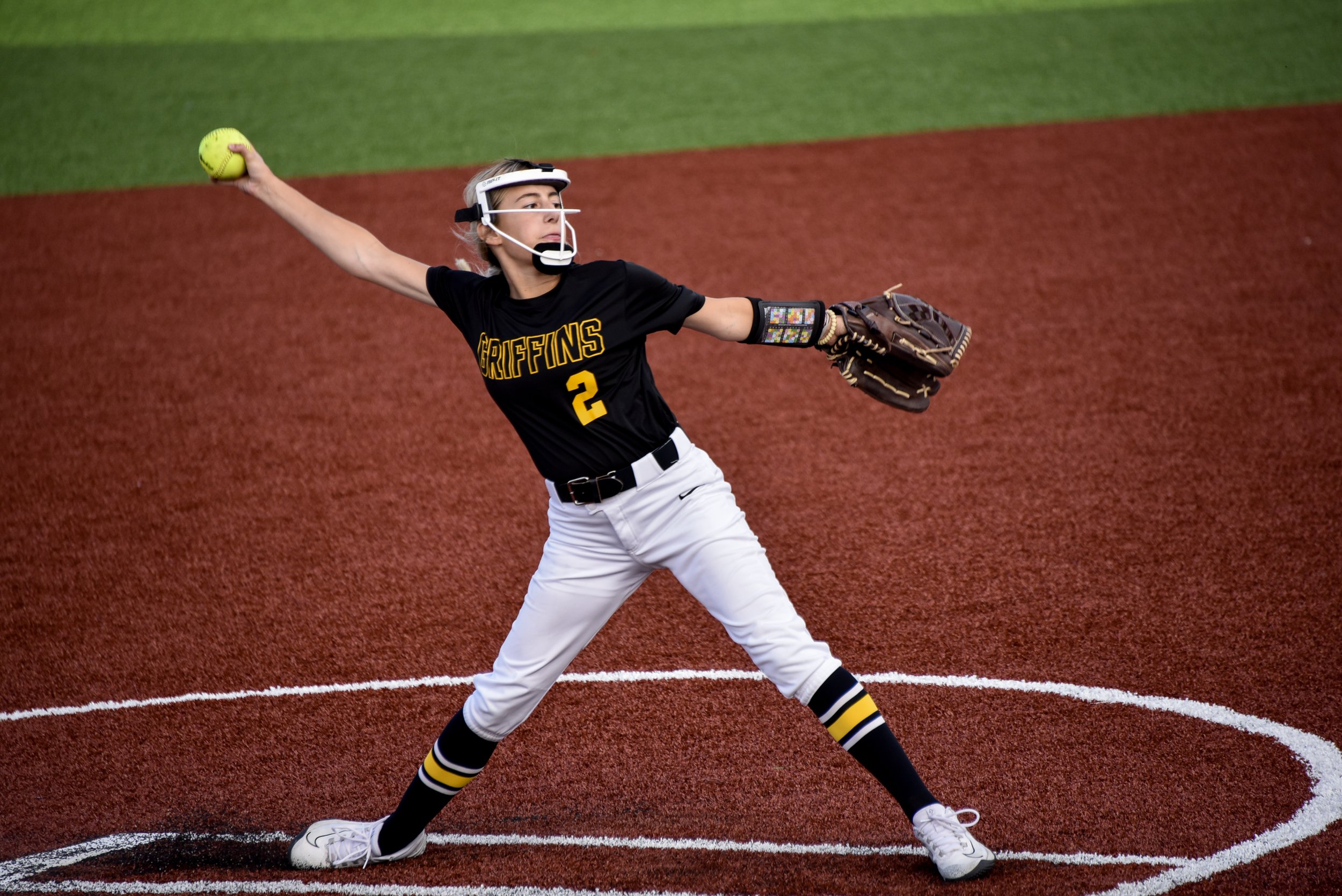 A female softball player in a black jersey with yellow lettering and the number 2 throws a yellow softball from the pitcher's mound on a red and green field.