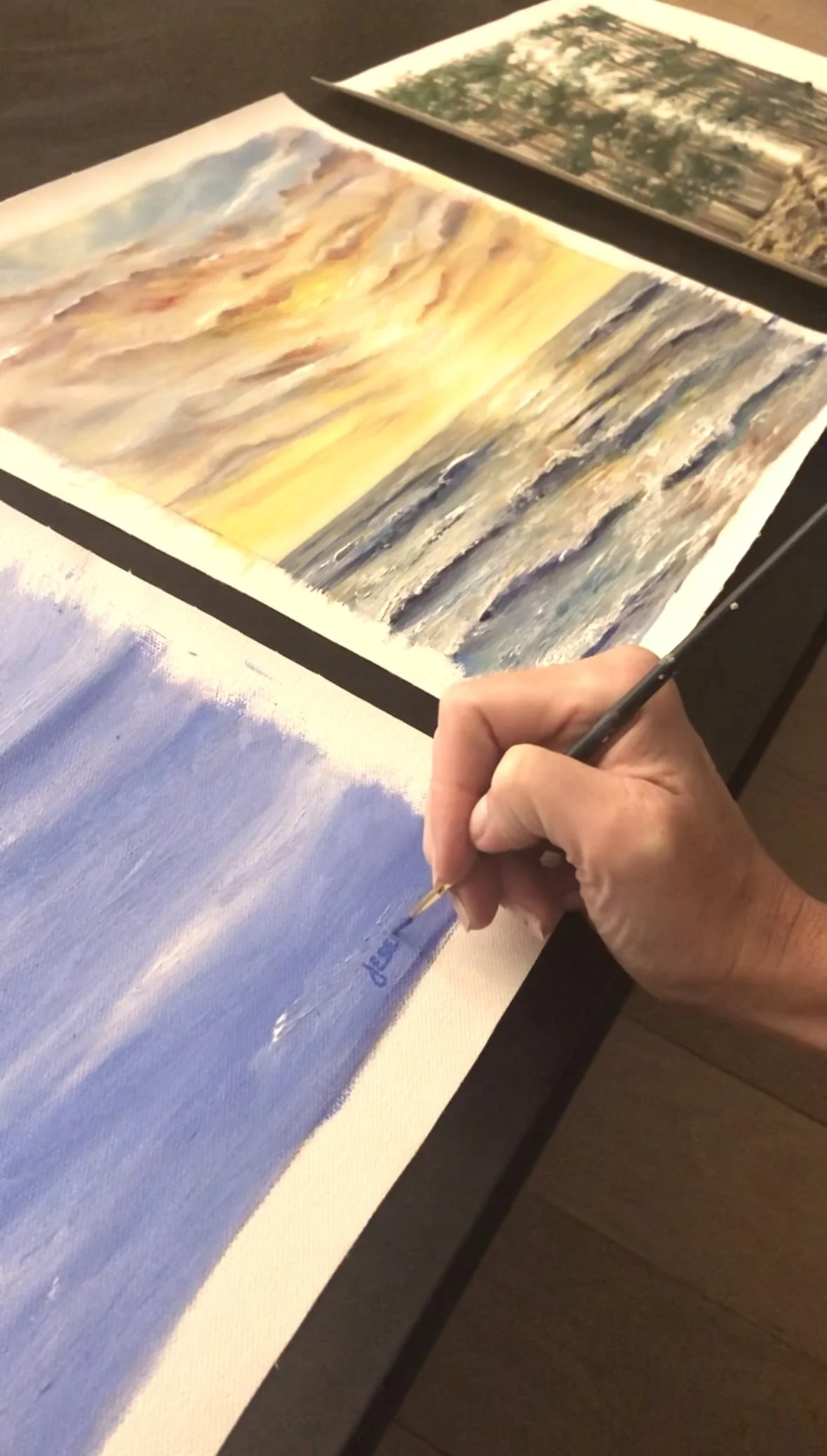 Hand painting a seascape on canvas with a fine brush, with finished ocean and mountain landscape paintings in the background.