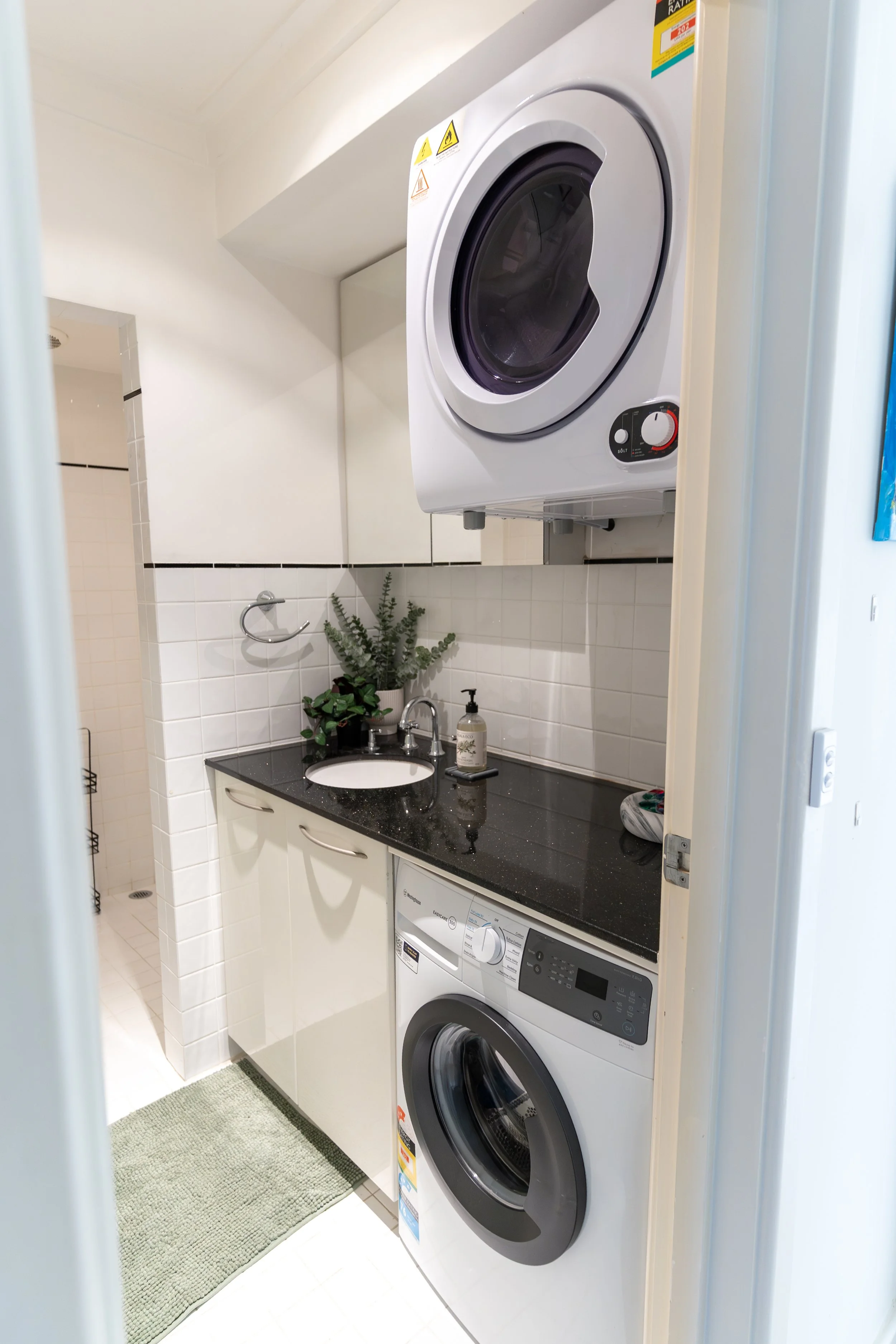 A laundry room with a washing machine beneath a beige countertop, a stainless steel sink with a faucet, and two potted plants on the counter. Above the counter, there's a wall-mounted white dryer. The room has white tiled walls and floor.