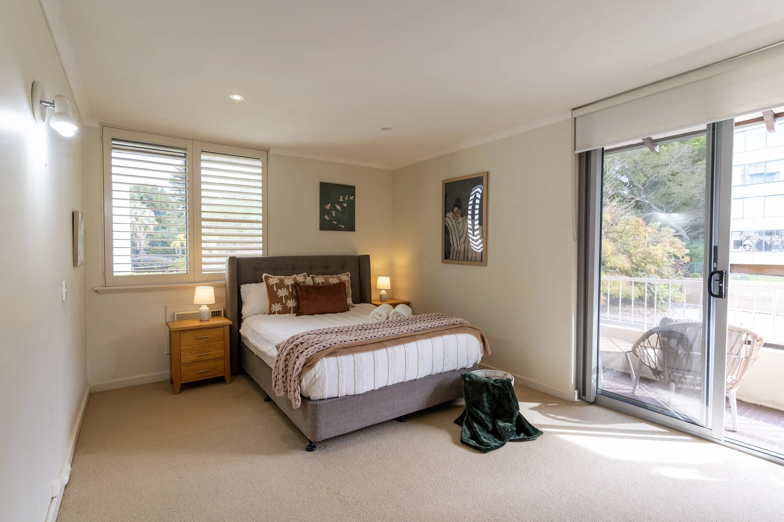 A neatly made bed with a brown headboard, two pillows, and a decorative knot pillow, flanked by two wooden nightstands with modern lamps. A piece of abstract art hangs above the bed, and a window allows natural light into the room.