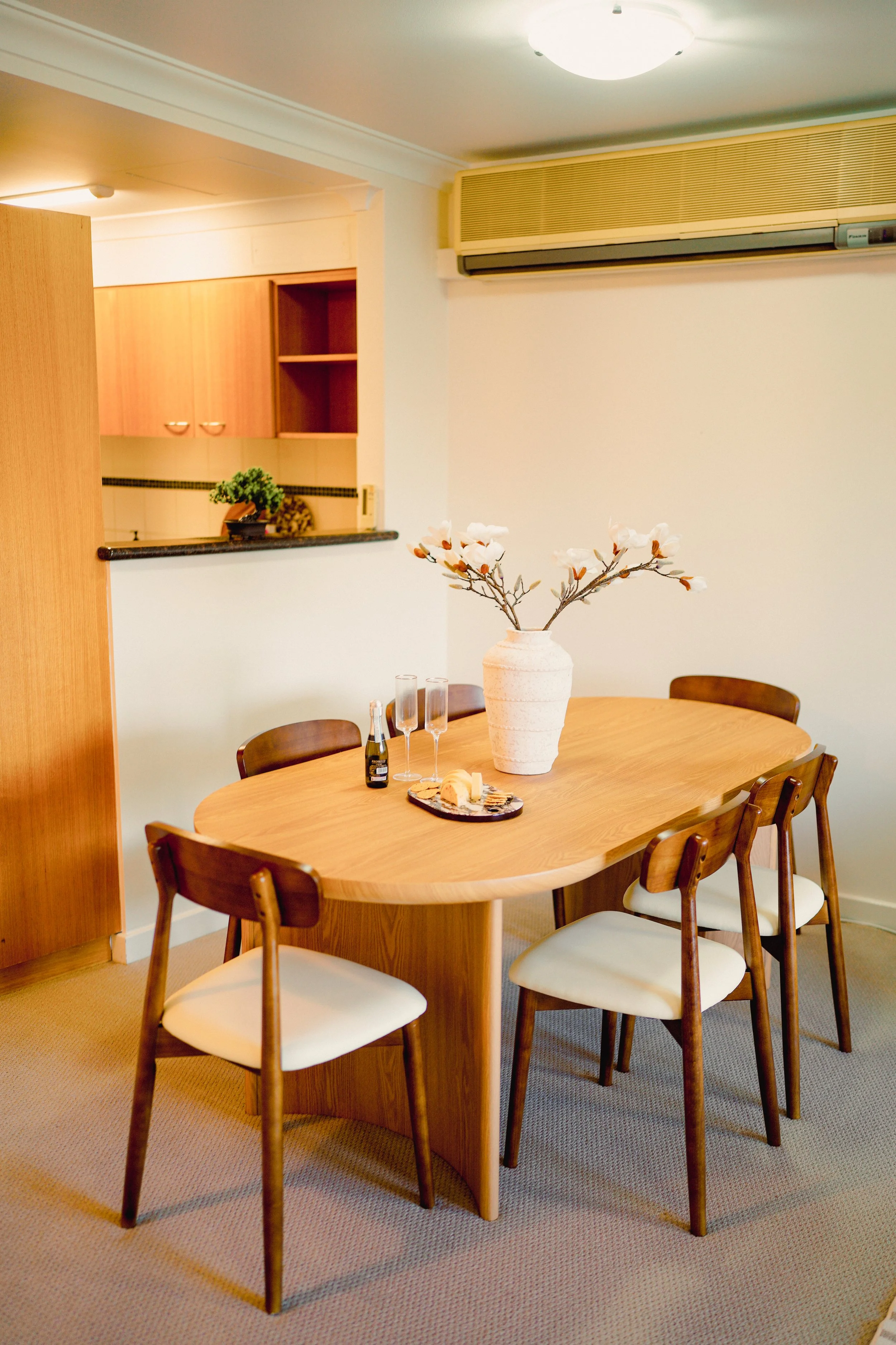 A round wooden dining table set with four place settings, each with a placemat, napkins, and silverware. A green and white striped vase with flowers is in the center. Four teal chairs are around the table, and a kitchen with white cabinets and a window are visible in the background.
