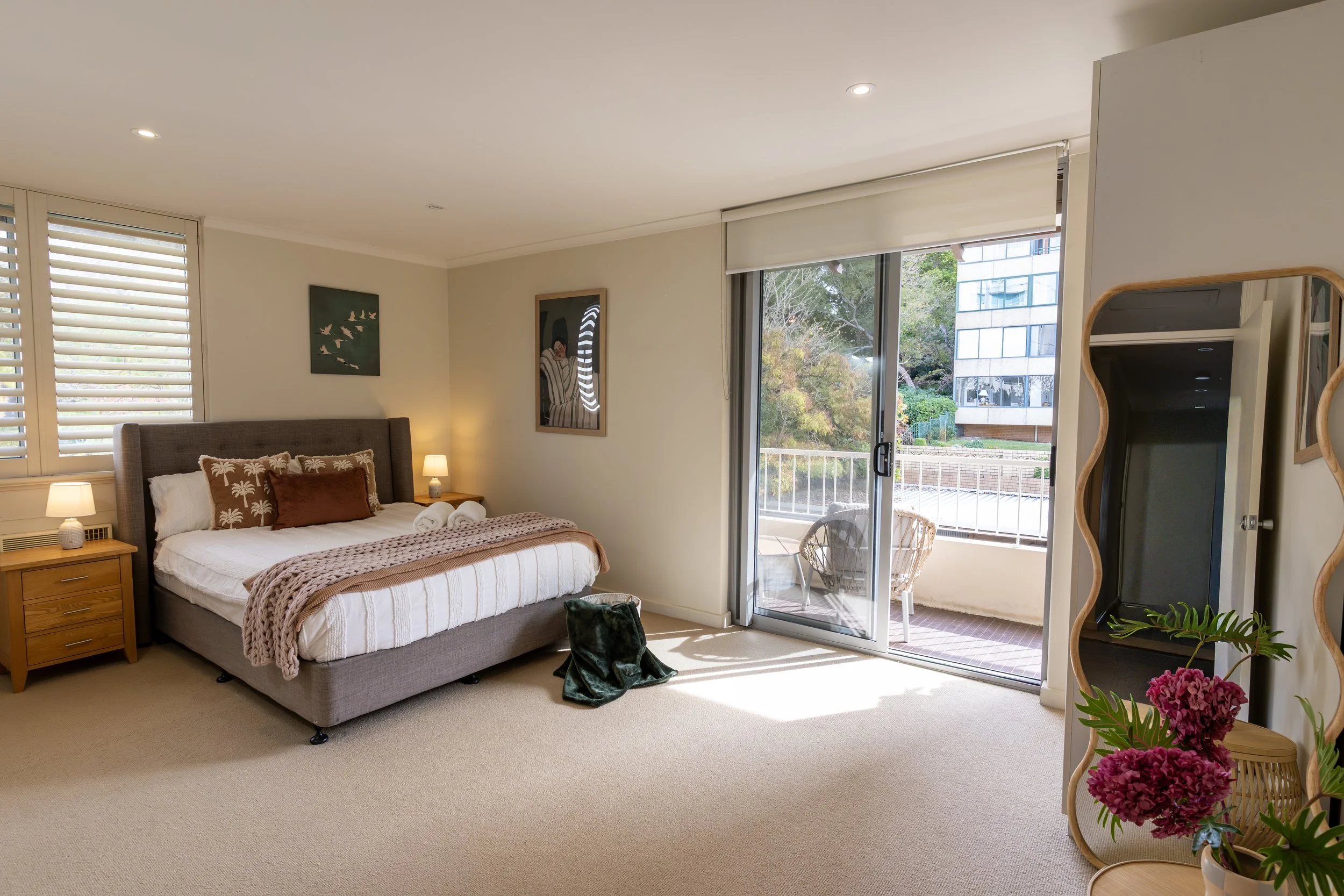 A neatly made bed with white embroidered bedding, a dark gray upholstered headboard, two green and white striped pillows, a towel folded into a decorative knot shape, and a dark wooden nightstand with a metallic lamp on each side, placed beside a window.