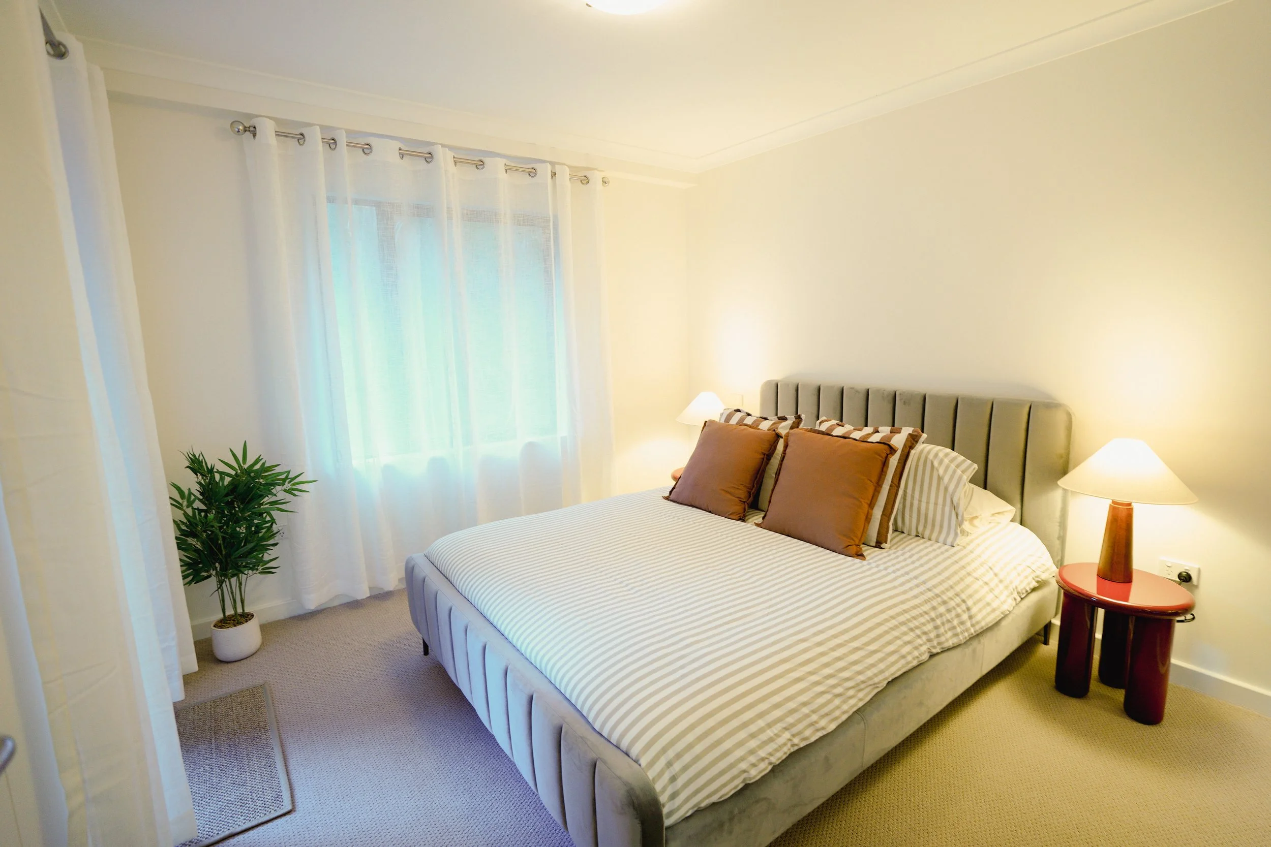 A neatly made bed with a gray upholstered headboard, white textured bedding, decorative pillows, and a brown blanket. Two wooden nightstands with small lamps are on either side of the bed. There is a colorful abstract painting above the headboard and a window to the right showing part of a building outside.