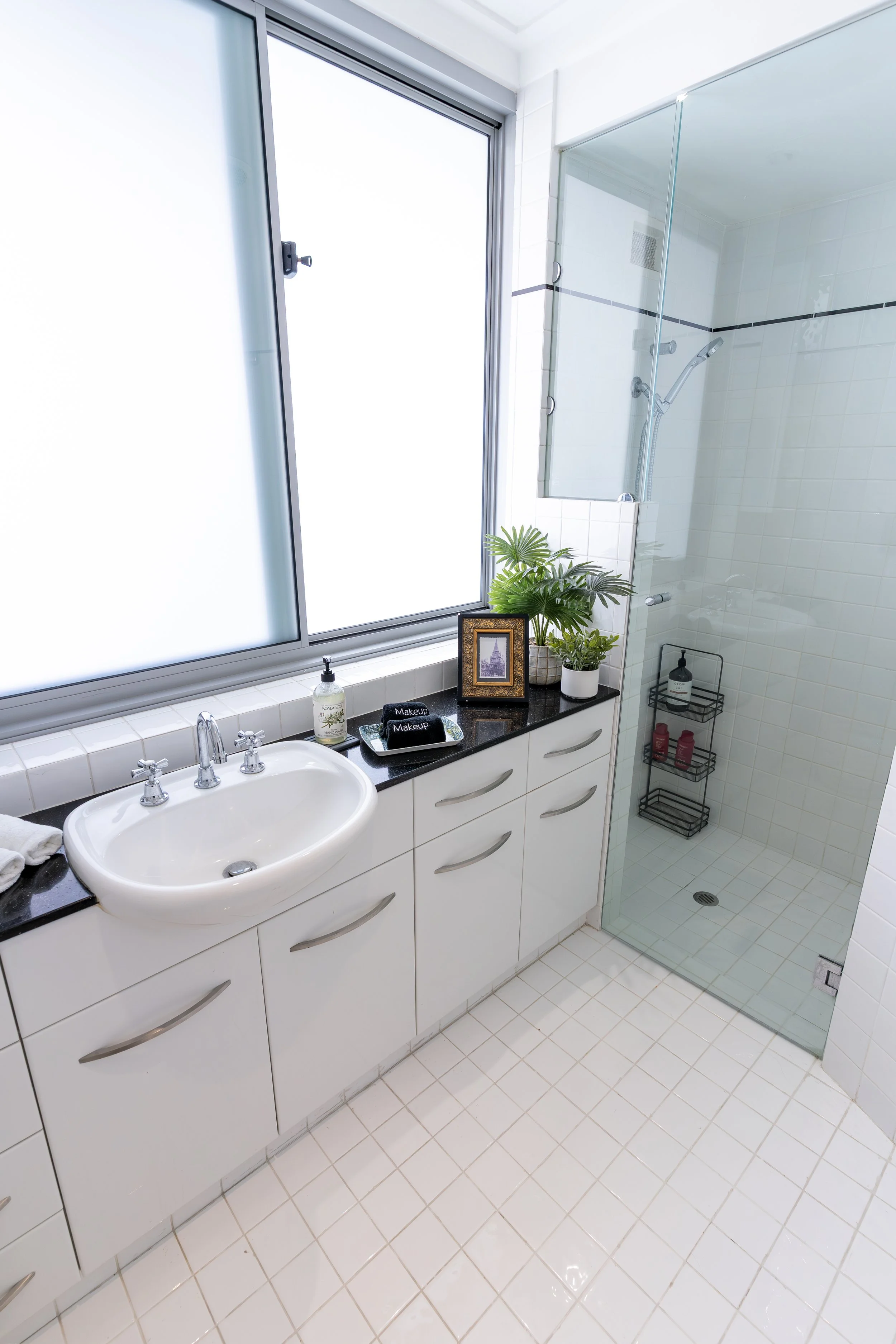 A modern bathroom with beige tiled flooring, a shower with glass doors, a white toilet, a vanity with a sink, a mirror, a potted plant, and laundry appliances visible in the reflection.