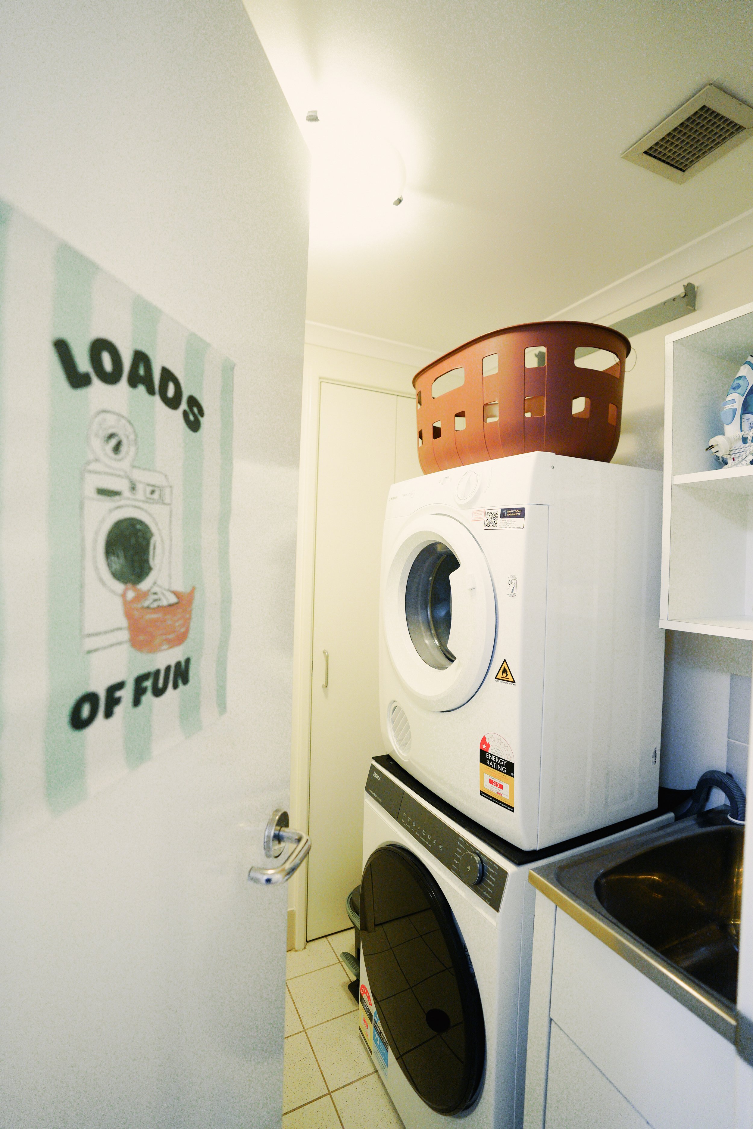 A laundry room with a washing machine beneath a beige countertop, a stainless steel sink with a faucet, and two potted plants on the counter. Above the counter, there's a wall-mounted white dryer. The room has white tiled walls and floor.