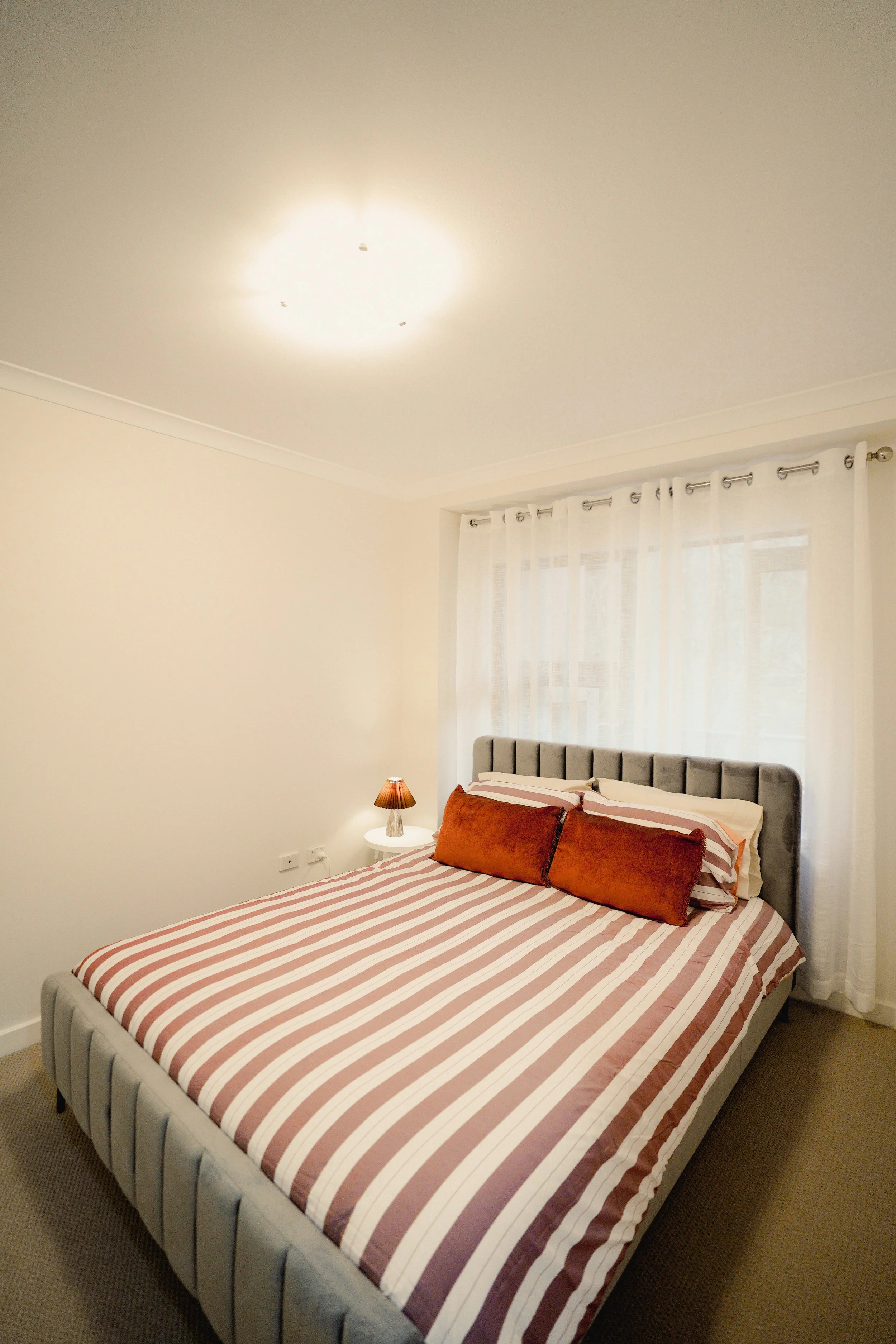 A neatly made bed with white embroidered bedding, a dark gray upholstered headboard, two green and white striped pillows, a towel folded into a decorative knot shape, and a dark wooden nightstand with a metallic lamp on each side, placed beside a window.