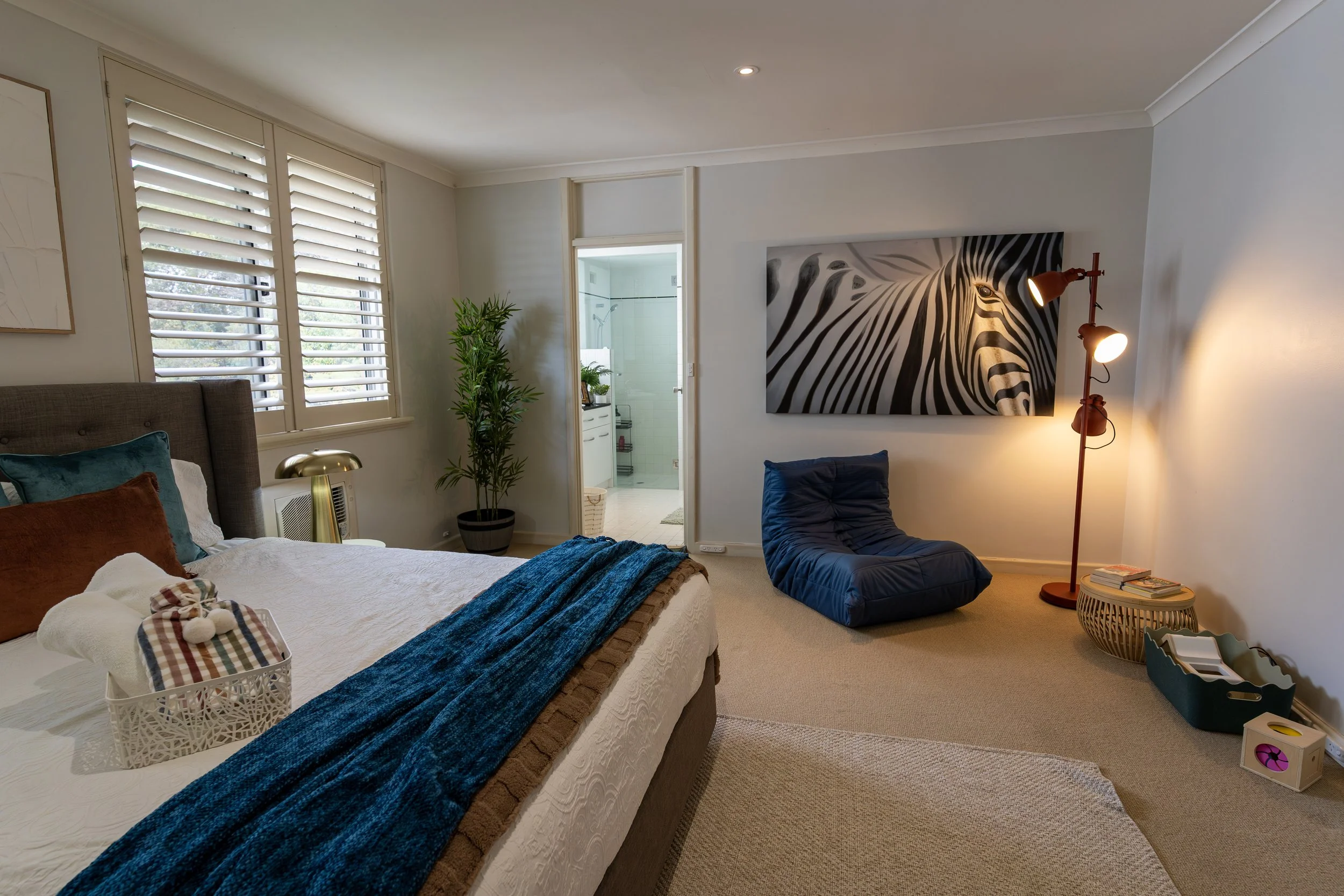 A neatly made bed with a gray upholstered headboard, white textured bedding, decorative pillows, and a brown blanket. Two wooden nightstands with small lamps are on either side of the bed. There is a colorful abstract painting above the headboard and a window to the right showing part of a building outside.