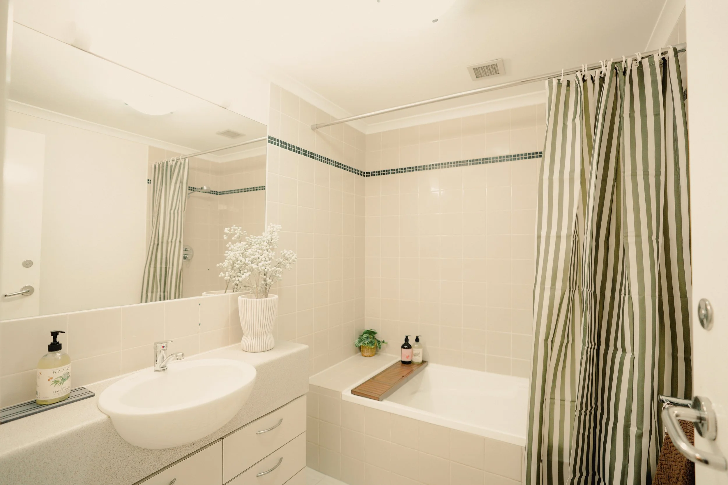 A modern bathroom with beige tiled flooring, a shower with glass doors, a white toilet, a vanity with a sink, a mirror, a potted plant, and laundry appliances visible in the reflection.