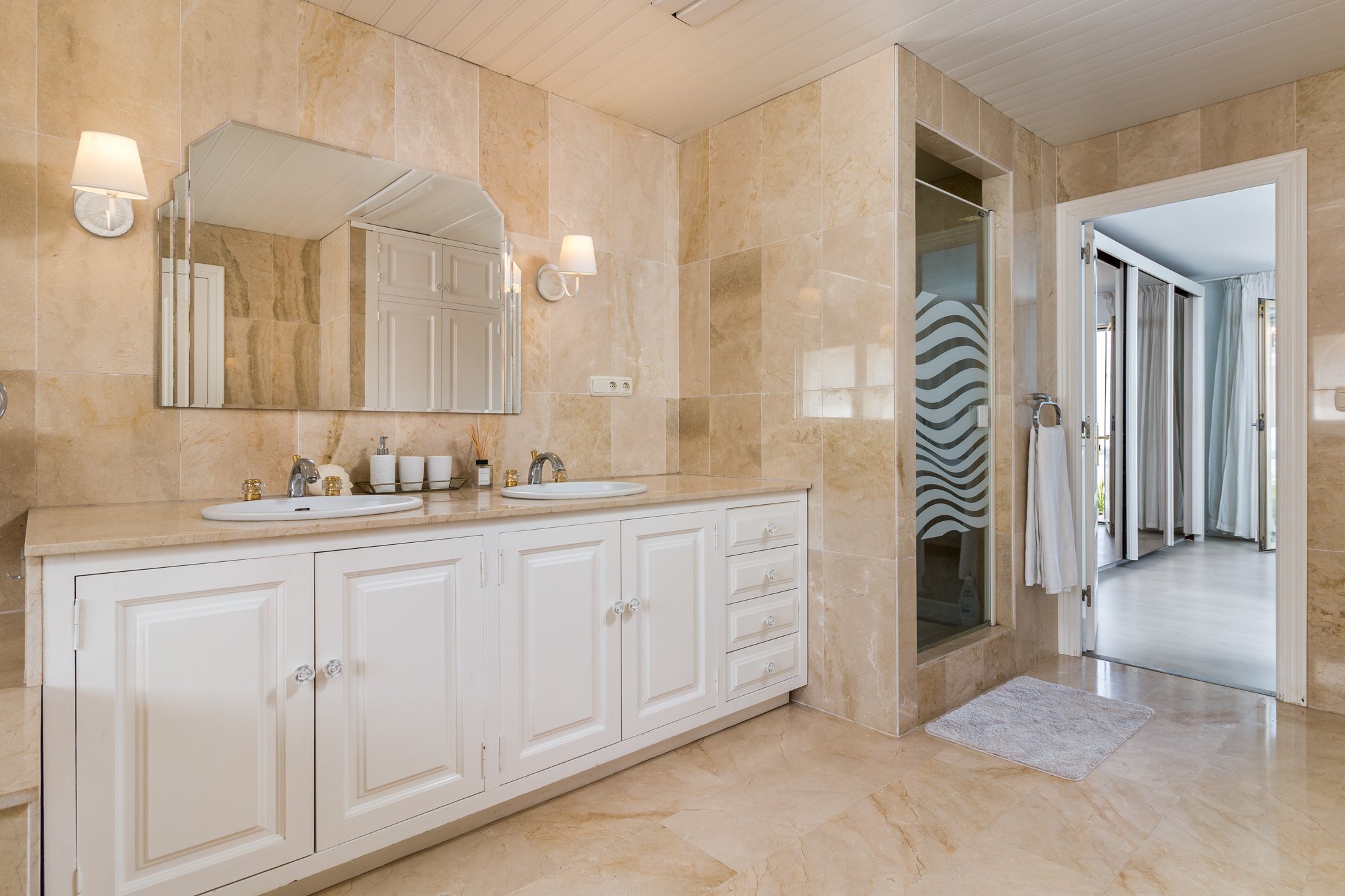 Luxury bathroom with marble tiles, double white vanity with sinks, large mirror, and glass shower door with wavy design.
