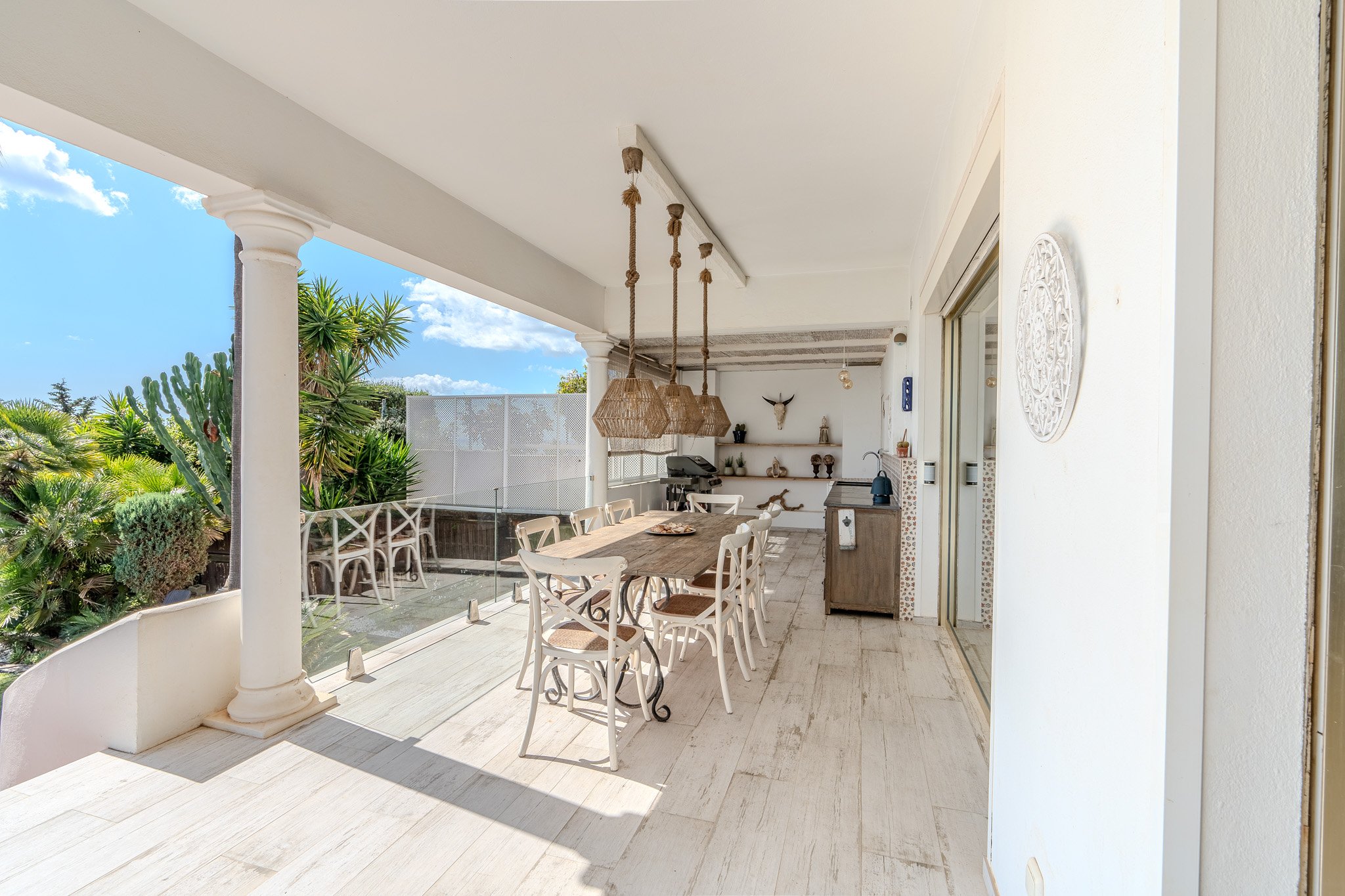 Outdoor patio with a wooden dining table and chairs, hanging woven lamps, decorative wall art, and tropical plants. Features include a grill and countertop.