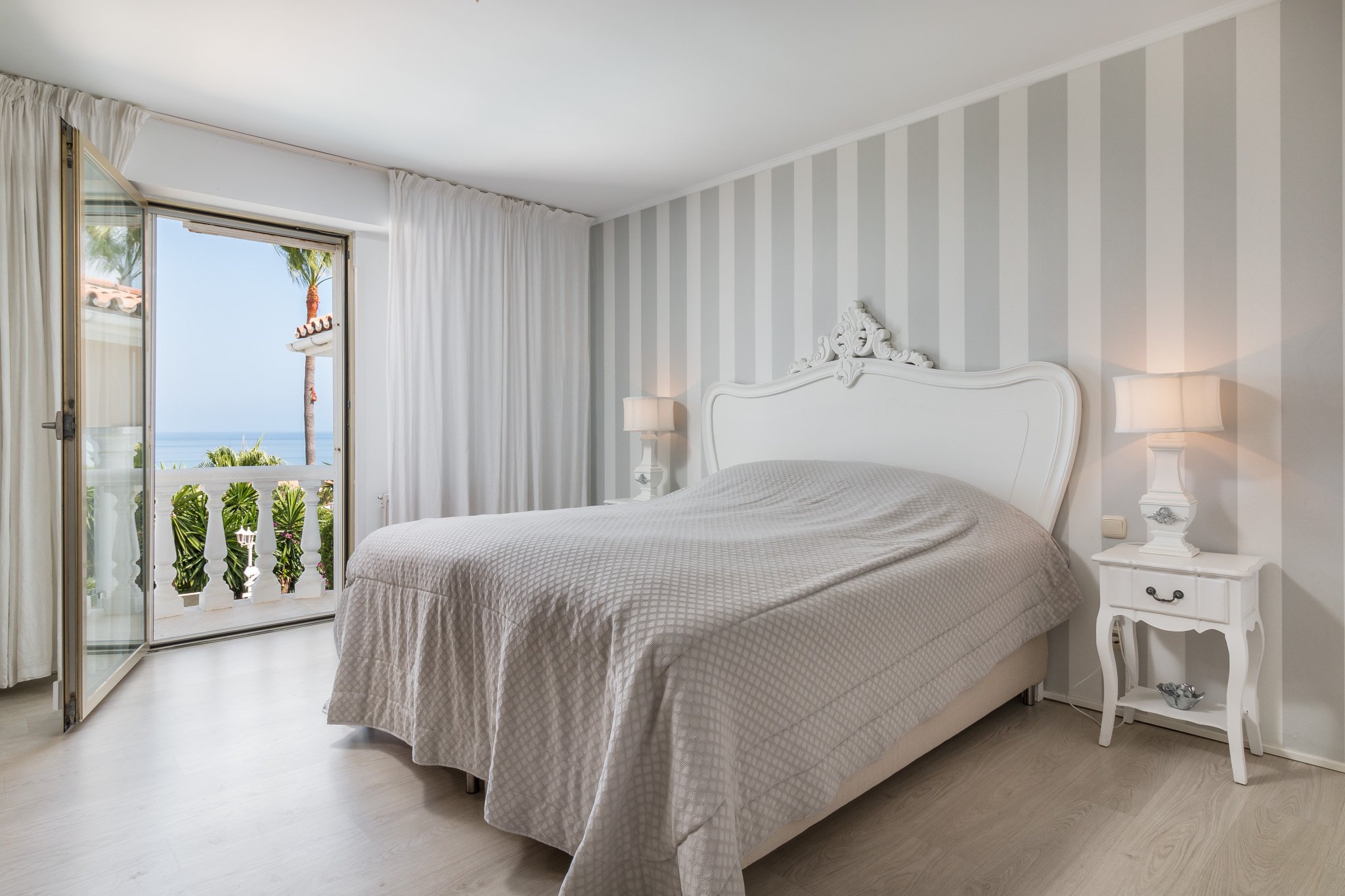 Bright bedroom with striped wall, large bed, white headboard, nightstands, lamps, open doors to balcony, ocean view.
