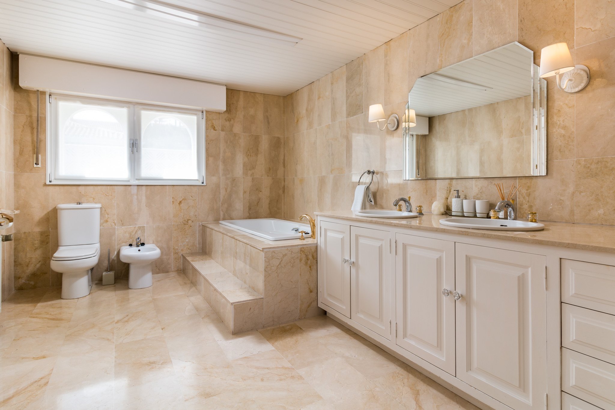 Luxury bathroom with beige marble tiles, white double vanity, large mirror, bathtub, toilet, and bidet.