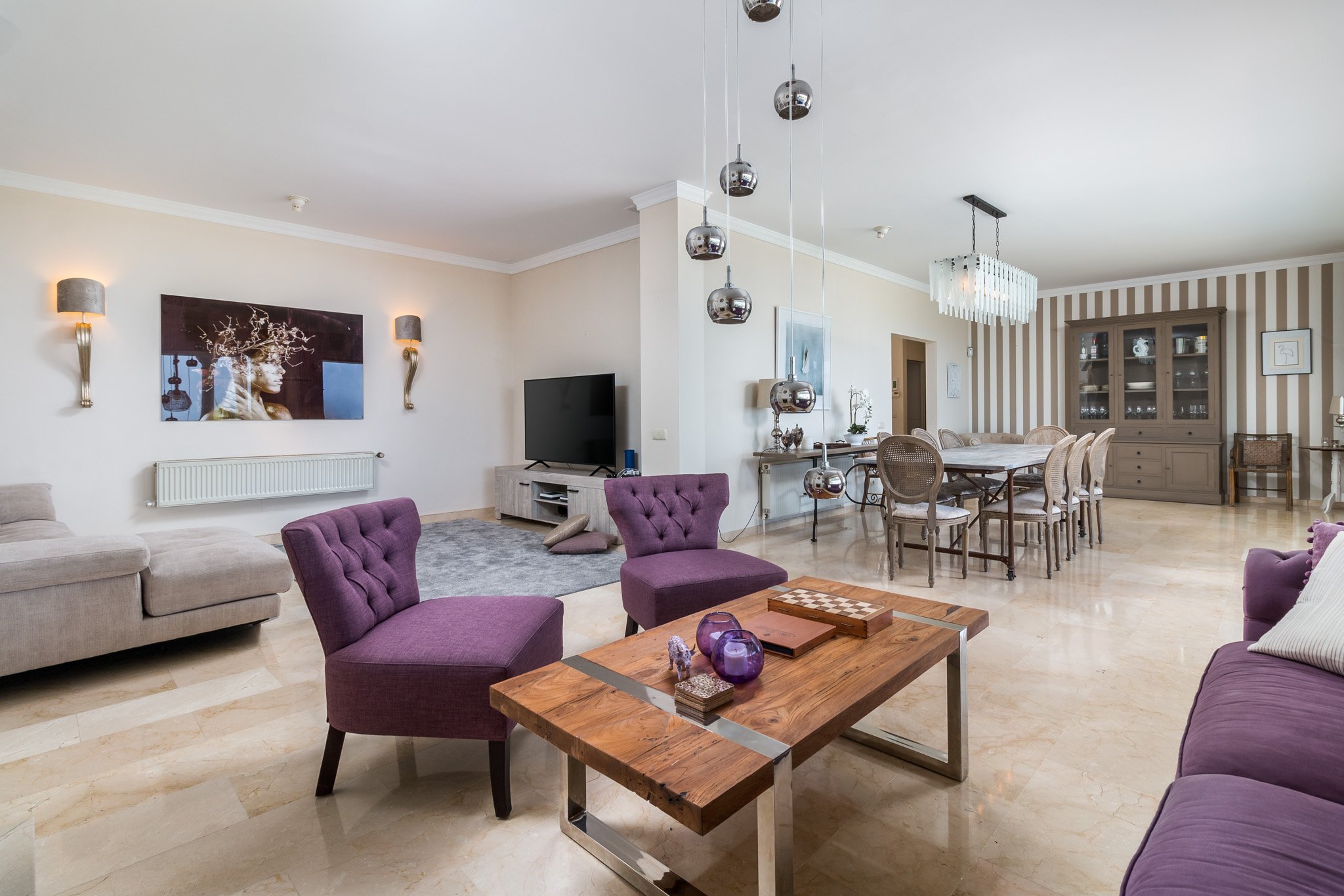 Modern living room with purple chairs, wooden coffee table, beige couch, and dining area in background. Decorative wall art and hanging pendant lights enhance the space. Large TV and cabinet near a window. Elegant and spacious design.