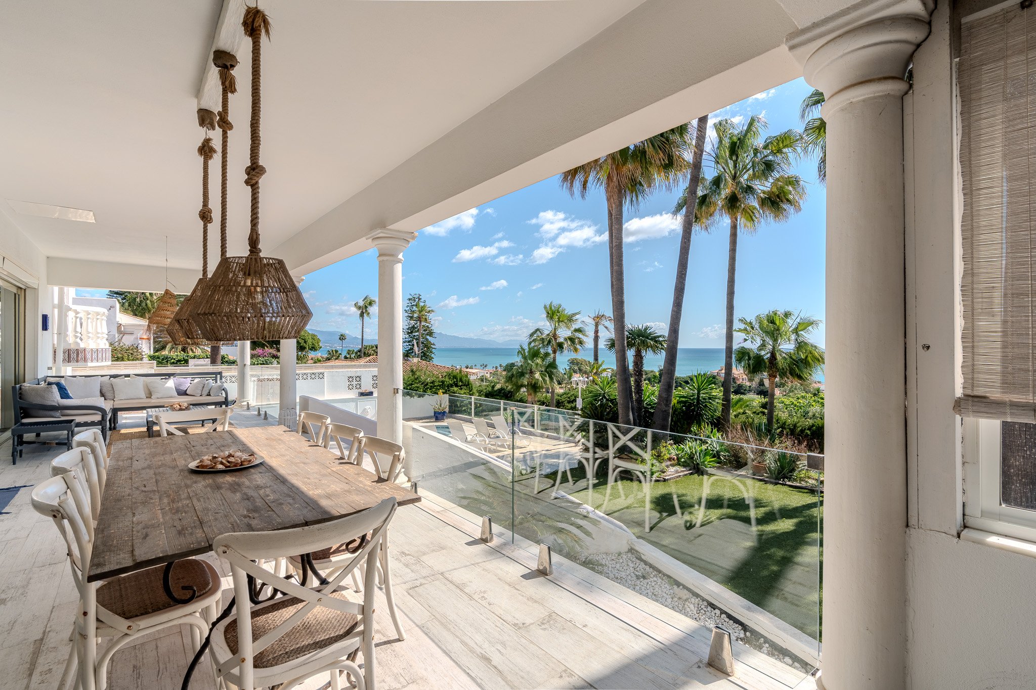 Outdoor patio with wooden dining table, wicker chairs, and hanging wicker lights overlooking lush garden and ocean view.