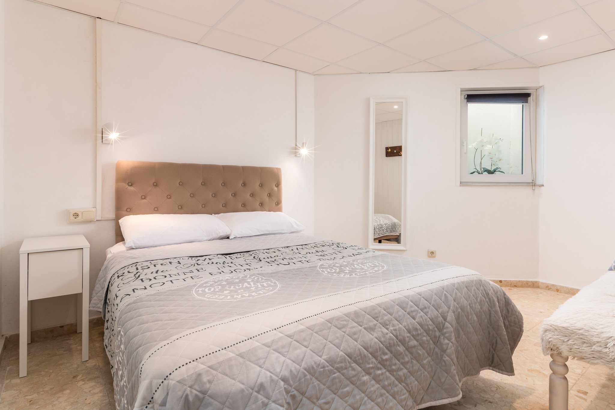 A bedroom with a beige upholstered headboard, white pillows, and a bed covered with a gray patterned quilt. A white nightstand is beside the bed. There's a wall mirror and a window with a floral curtain. The room has beige tiled flooring.