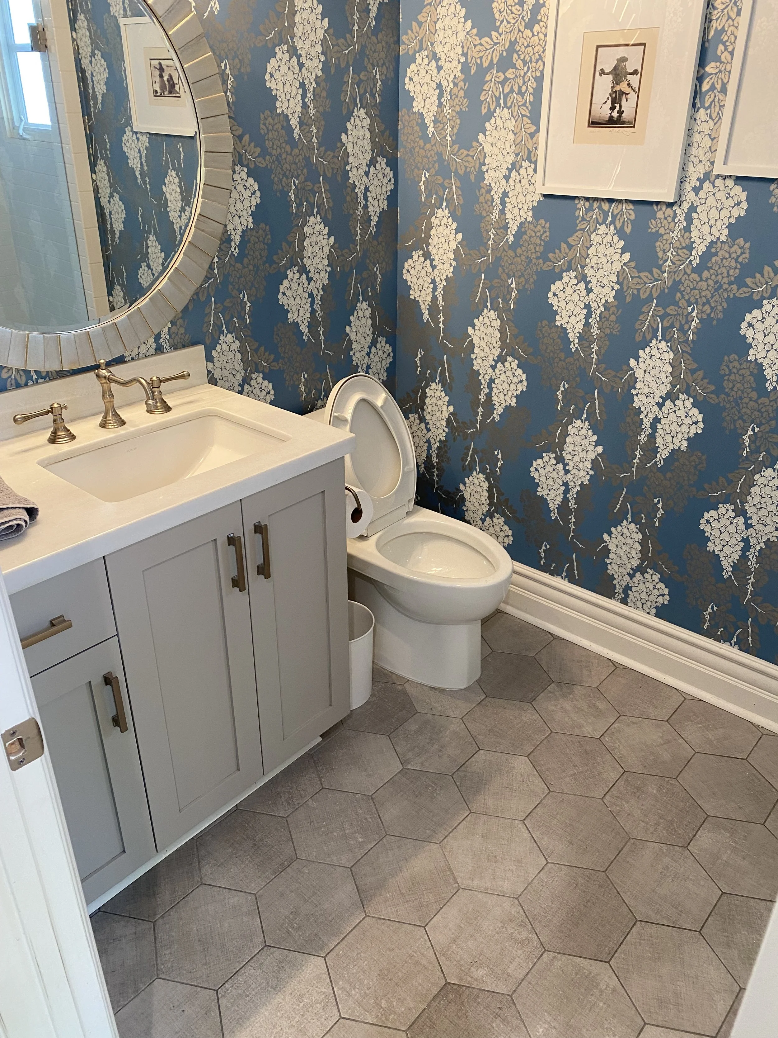 A bathroom with a white vanity and sink, a round mirror, a toilet, blue floral wallpaper, framed pictures, and hexagonal gray floor tiles.
