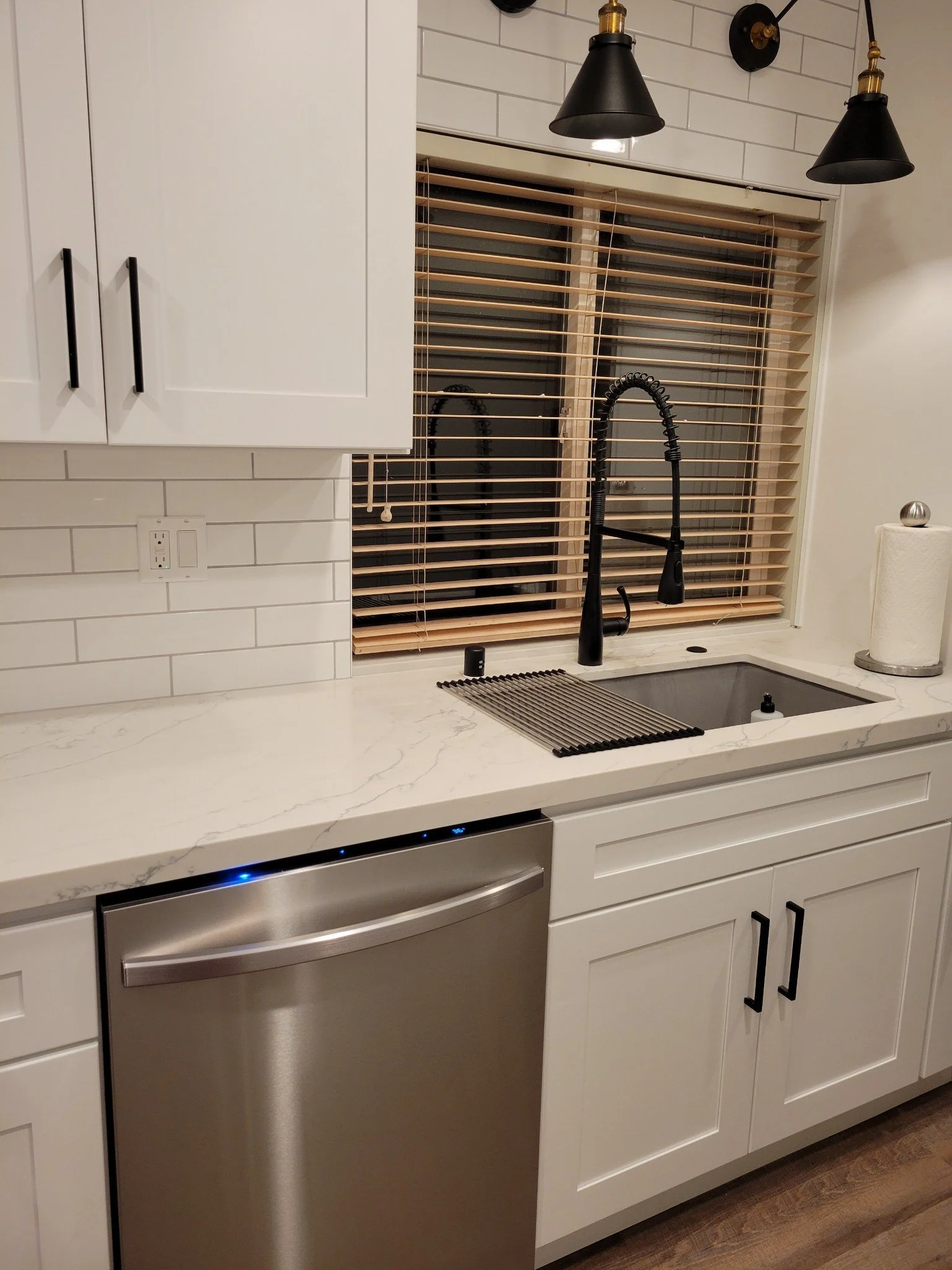 Modern kitchen with white cabinets, a marble countertop, a stainless steel dishwasher, and black fixtures including a faucet. There are wooden blinds on the window and two black pendant lights hanging above.