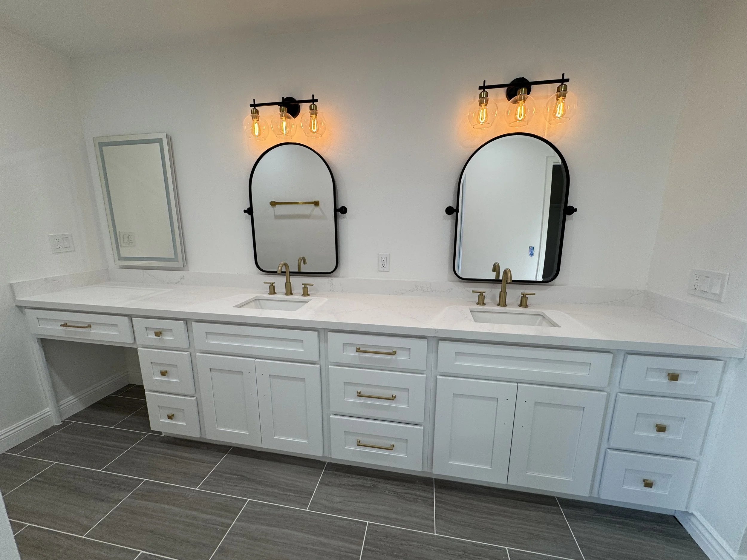 A modern white bathroom vanity with two sinks, gold faucets, and two oval mirrors above each sink. There are gold and black light fixtures with Edison bulbs above each mirror. The vanity has multiple drawers and cabinets with gold handles, and a marb