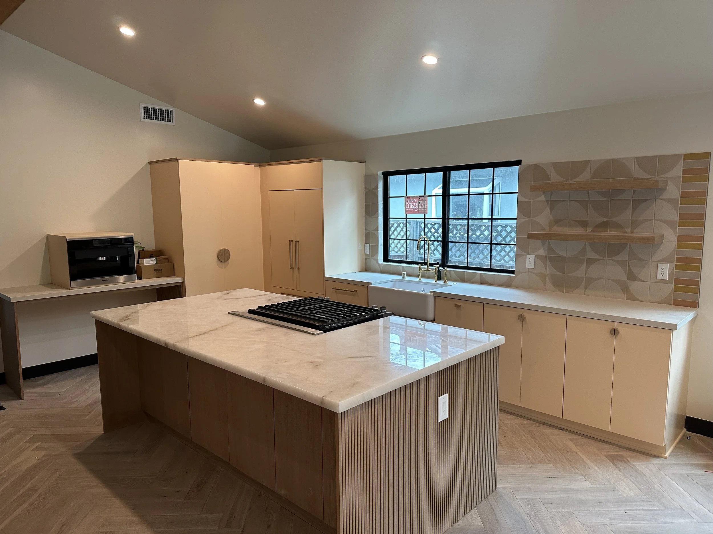 Modern kitchen with beige cabinets, wooden island with marble countertop, black stovetop, white farmhouse sink, window with black frame showing outdoor view, geometric patterned backsplash, open shelving, and wood flooring.