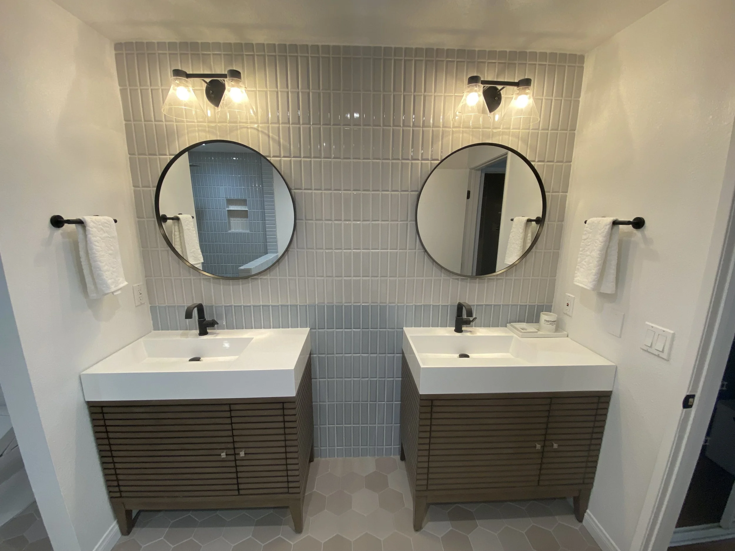 Bathroom with two white sinks, black faucets, round mirrors, black light fixtures, and beige tile walls.