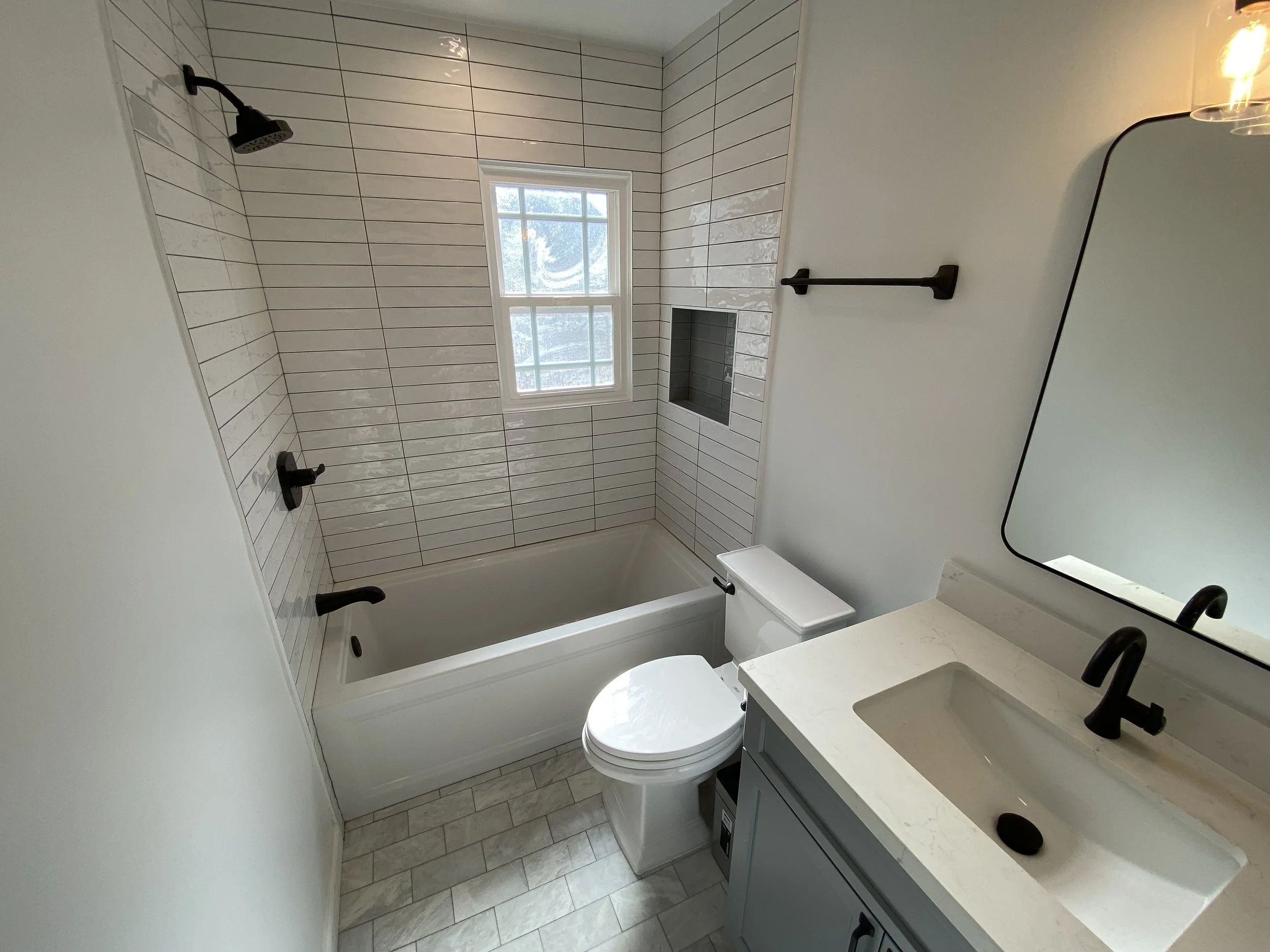 Small bathroom with white tiled walls, a bathtub with black fixtures, a small window above the tub, a toilet, a white vanity with a black faucet, a large mirror, and a black towel bar.