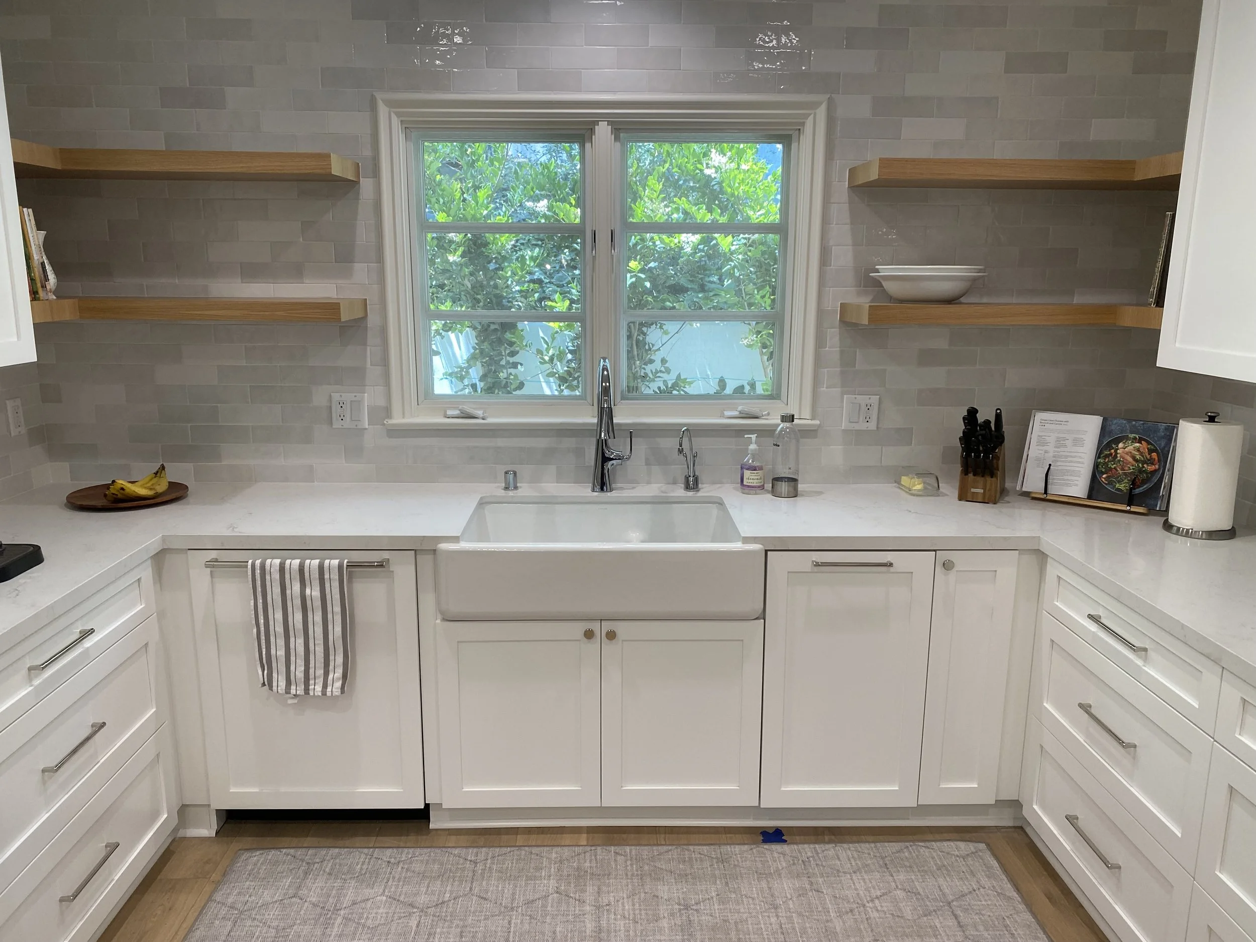 A clean kitchen with white cabinets, a double window above a farmhouse sink, open wooden shelves, banana on a plate, kitchen utensils, cookbook, paper towel roll, and some kitchen items on the countertop.
