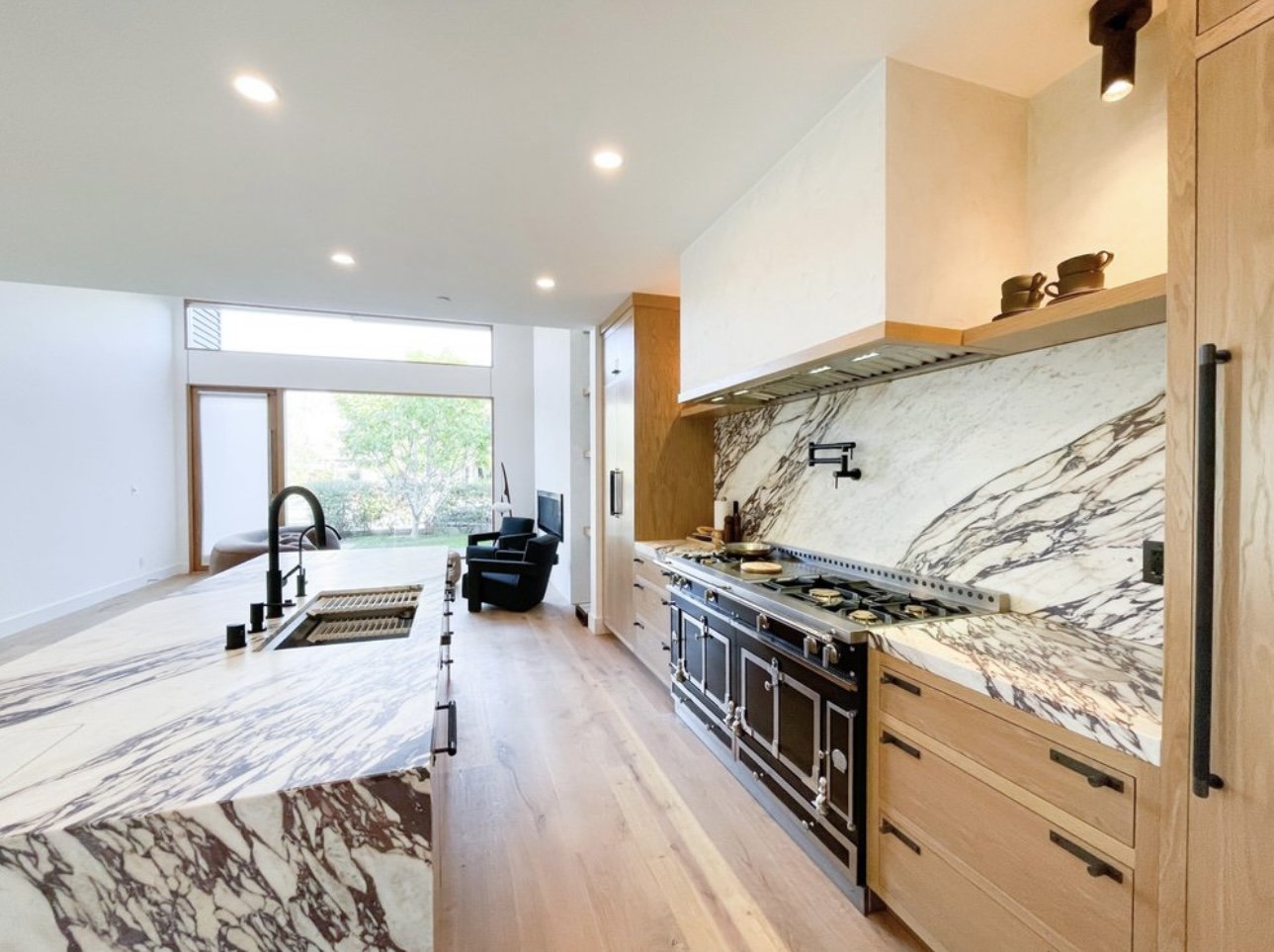 Modern kitchen with marble countertops and backsplash, black stove, wooden cabinets, open layout, and a view of a patio and outdoor greenery.