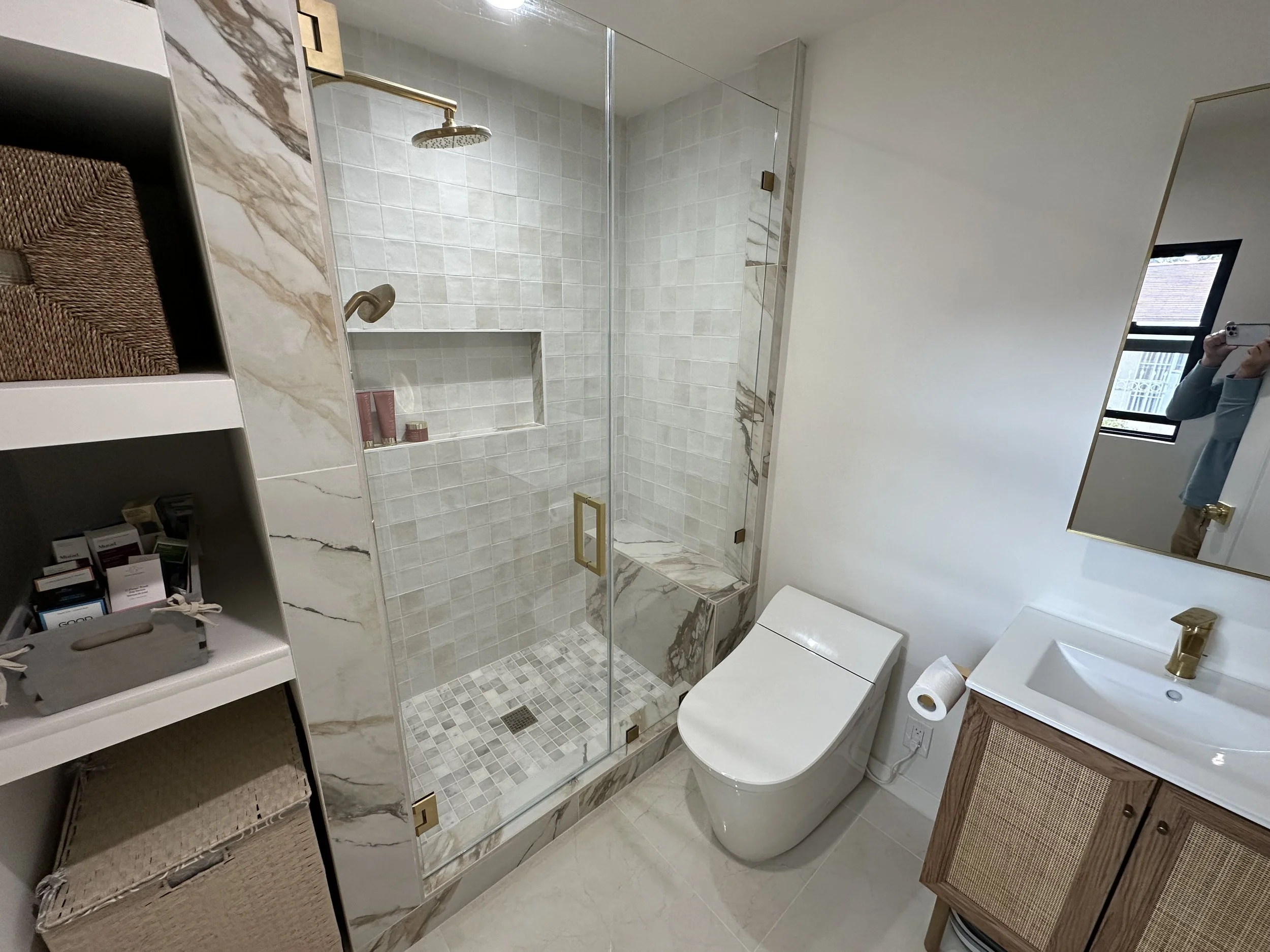 Modern bathroom with a glass-enclosed shower, a wall-mounted toilet, a white sink with a gold faucet, a mirror, and shelving with baskets.