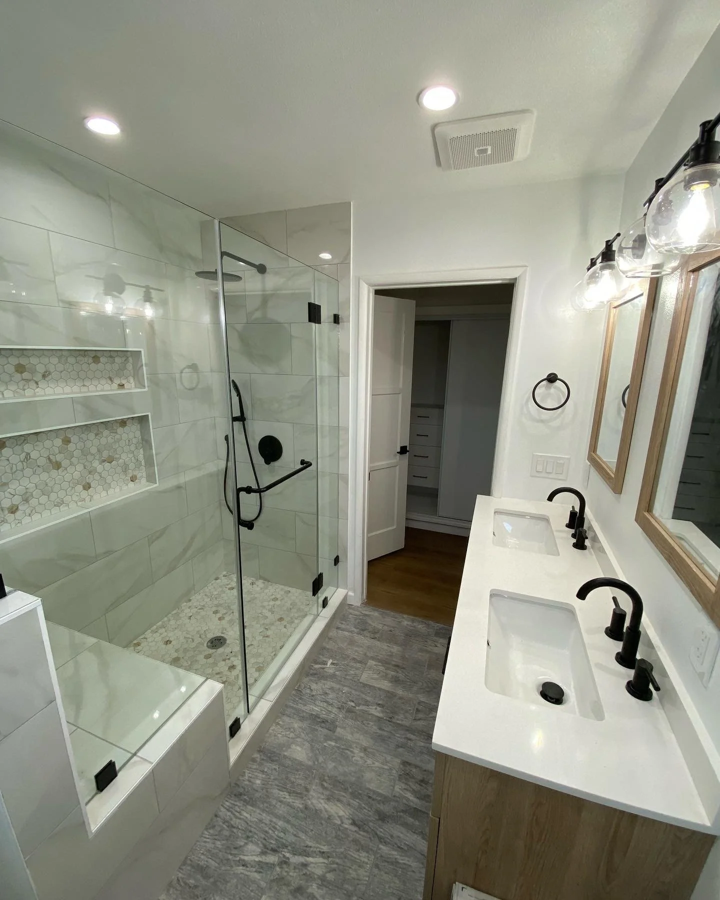 Modern bathroom with double vanity sinks featuring black fixtures, framed mirrors, glass enclosed shower with built-in shelves, and gray wood-look tile flooring.