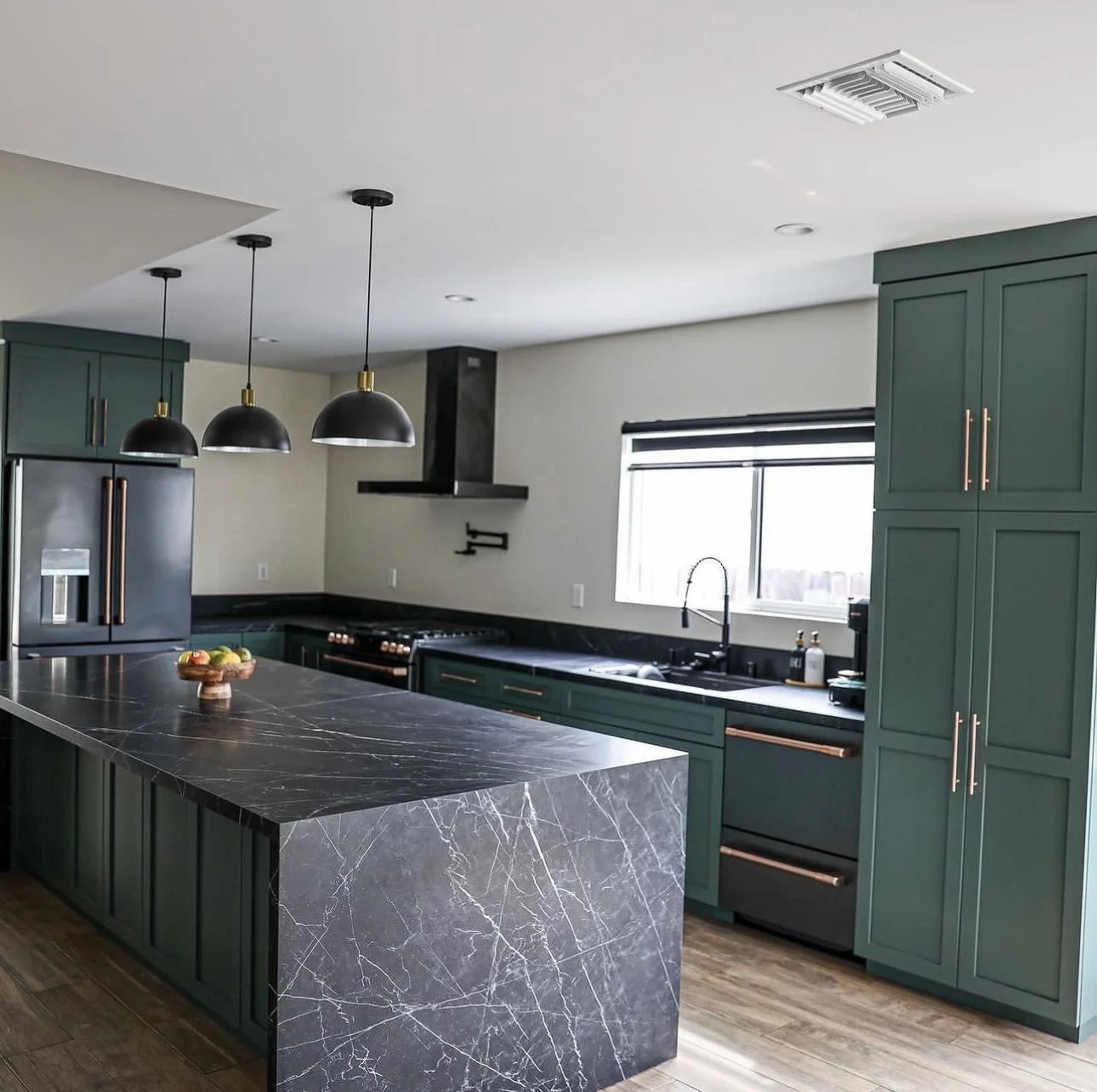 Modern kitchen with green cabinets, black marble countertop island, black appliances, and three black pendant lights.