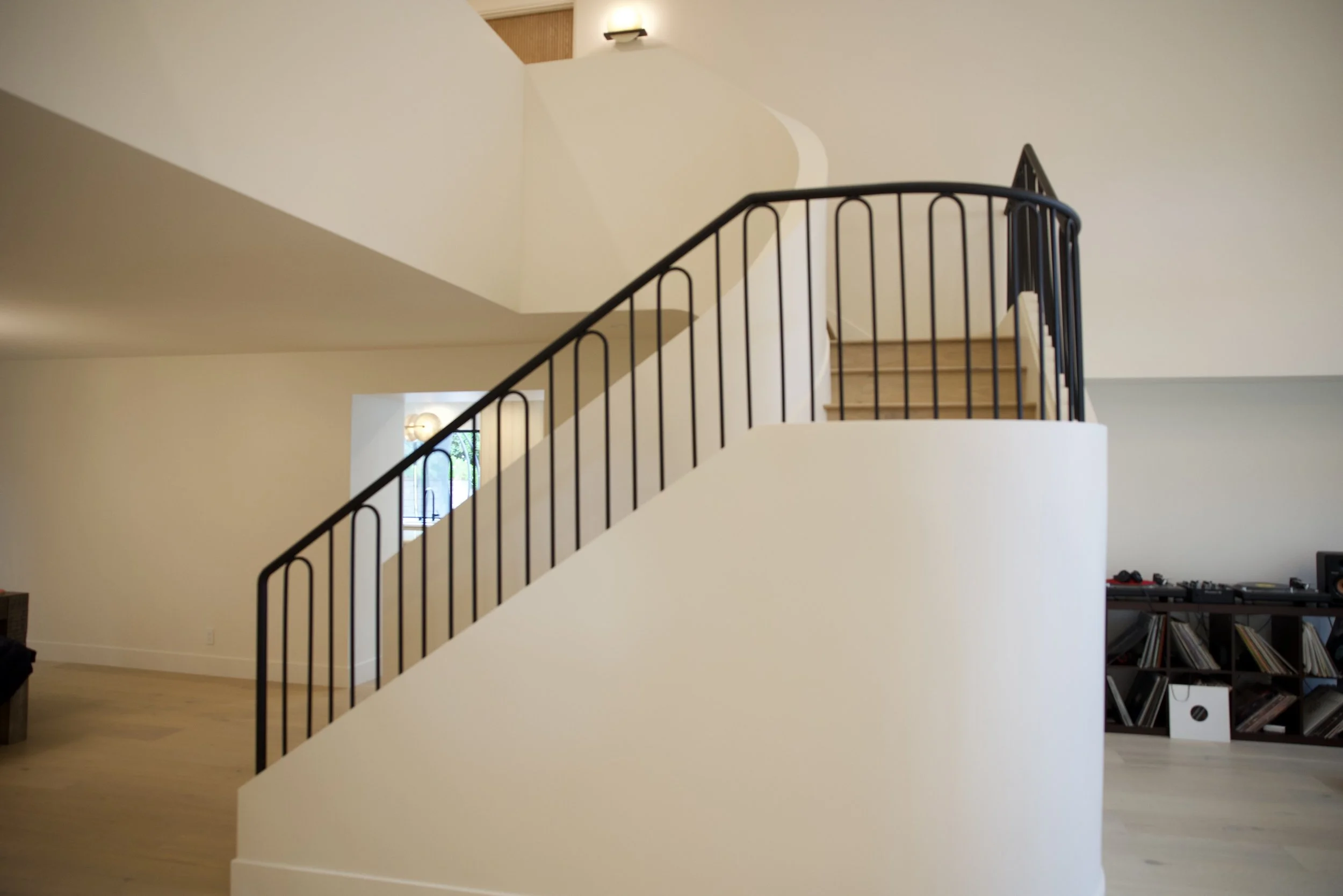 Interior view of a modern home with a curved staircase featuring black metal railing, leading to an upper floor, with a window and wall-mounted light fixture in the background.