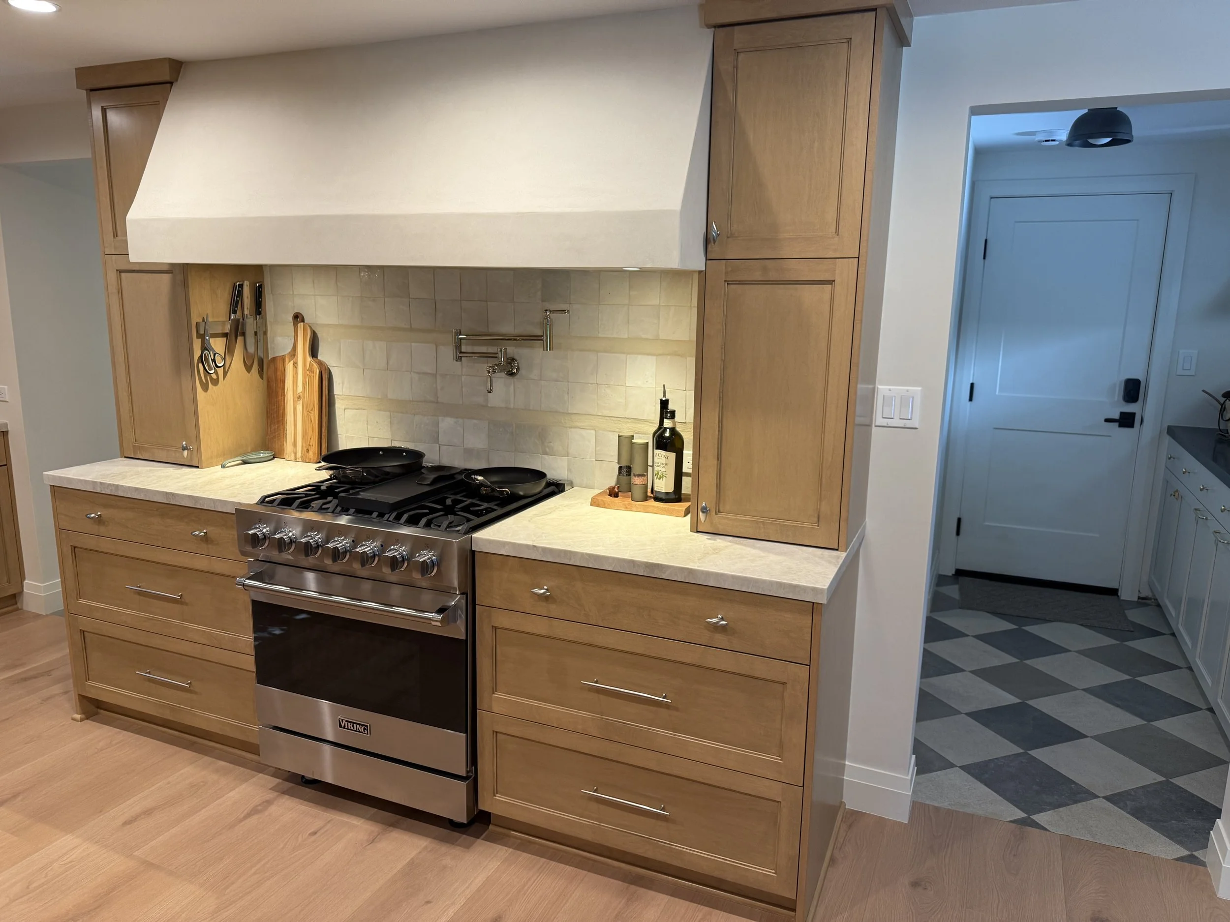 Kitchen with wooden cabinets, a stove, cutting boards, knives, bottles of oil or vinegar, and a backsplash.