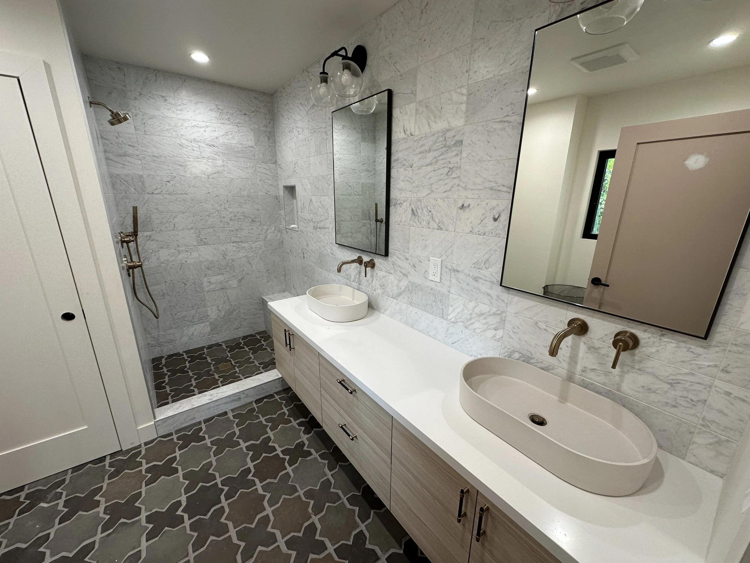 Modern bathroom with double vanity, oval vessel sinks, brass fixtures, large mirrors, tiled walk-in shower with niche, and patterned floor tiles.