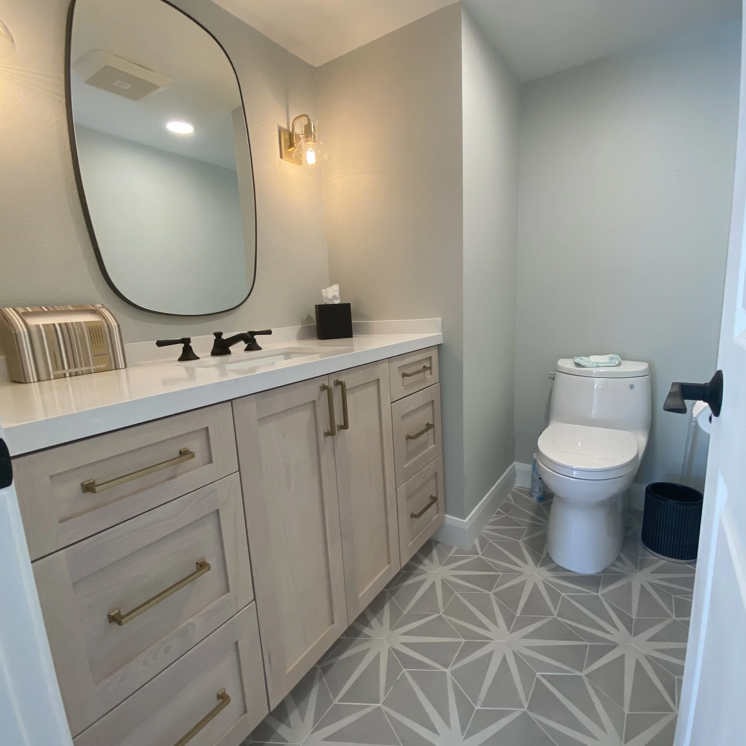 Modern bathroom with vanity and mirror on the left, toilet on the right, gray geometric floor tiles, and neutral-colored walls.